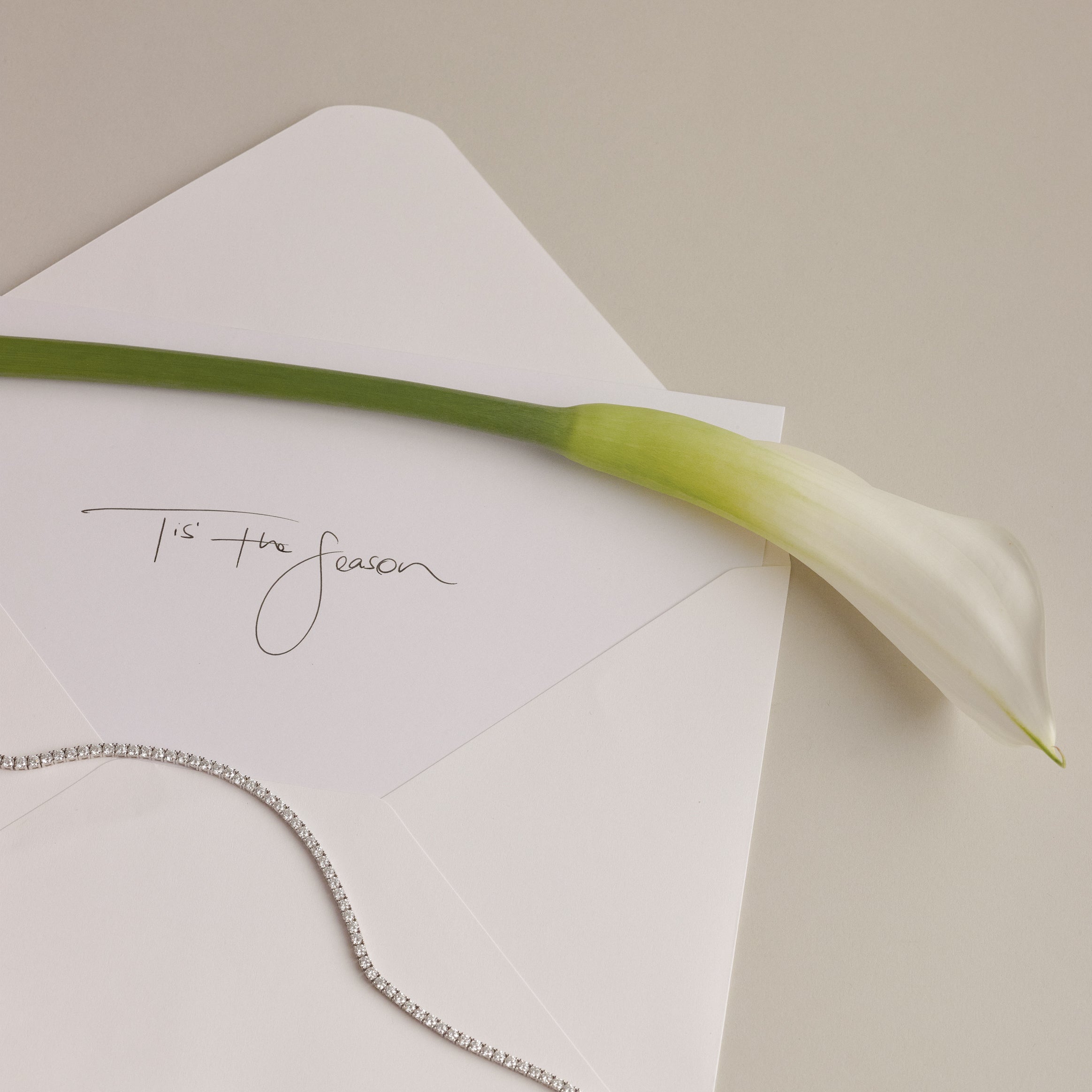 A beautiful cream envelope with a letter that writes 'Tis' the season', a tennis necklace is draped over the letter.