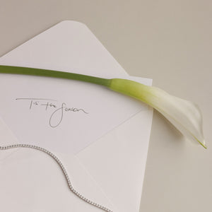 A beautiful cream envelope with a letter that writes 'Tis' the season', a tennis necklace is draped over the letter.