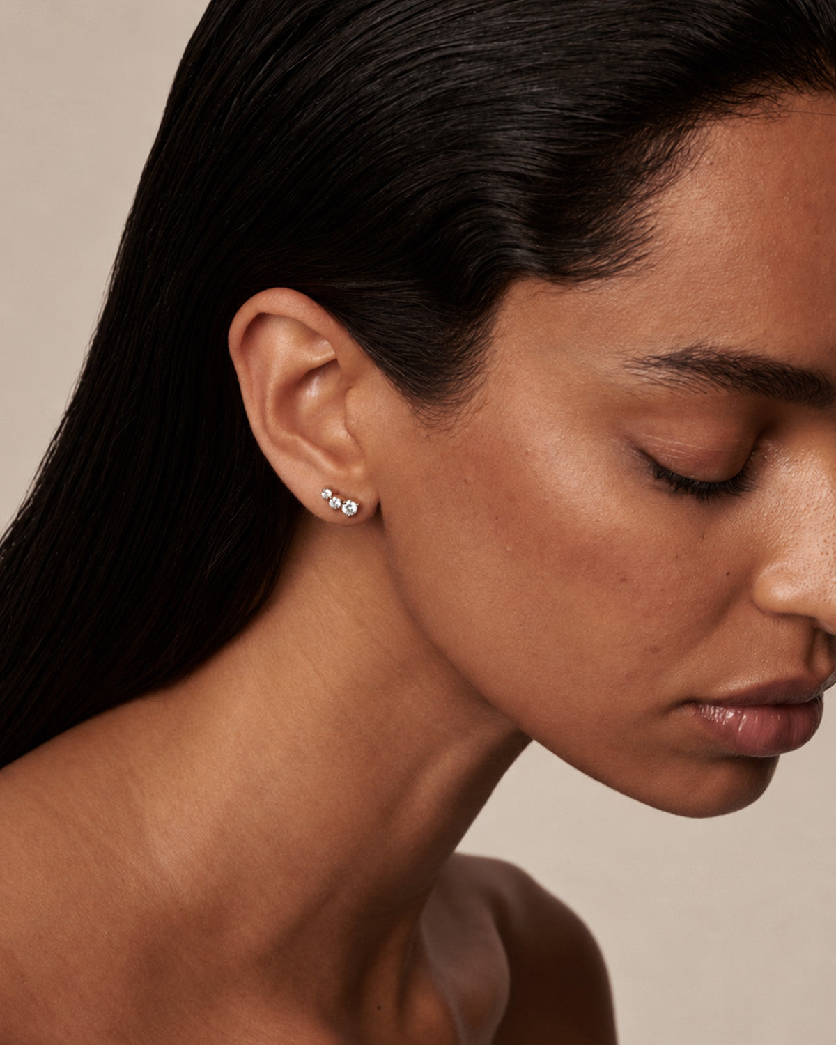 A close-up of a woman with dark hair wearing a small cluster stud earring with clear diamonds against a warm beige background.