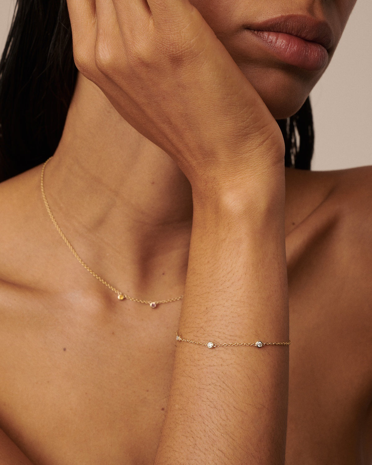 A close-up of a model's neck, shoulder, and arm wearing delicate gold jewelry, including a thin chain necklace and a matching bracelet with small bead and stone accents.