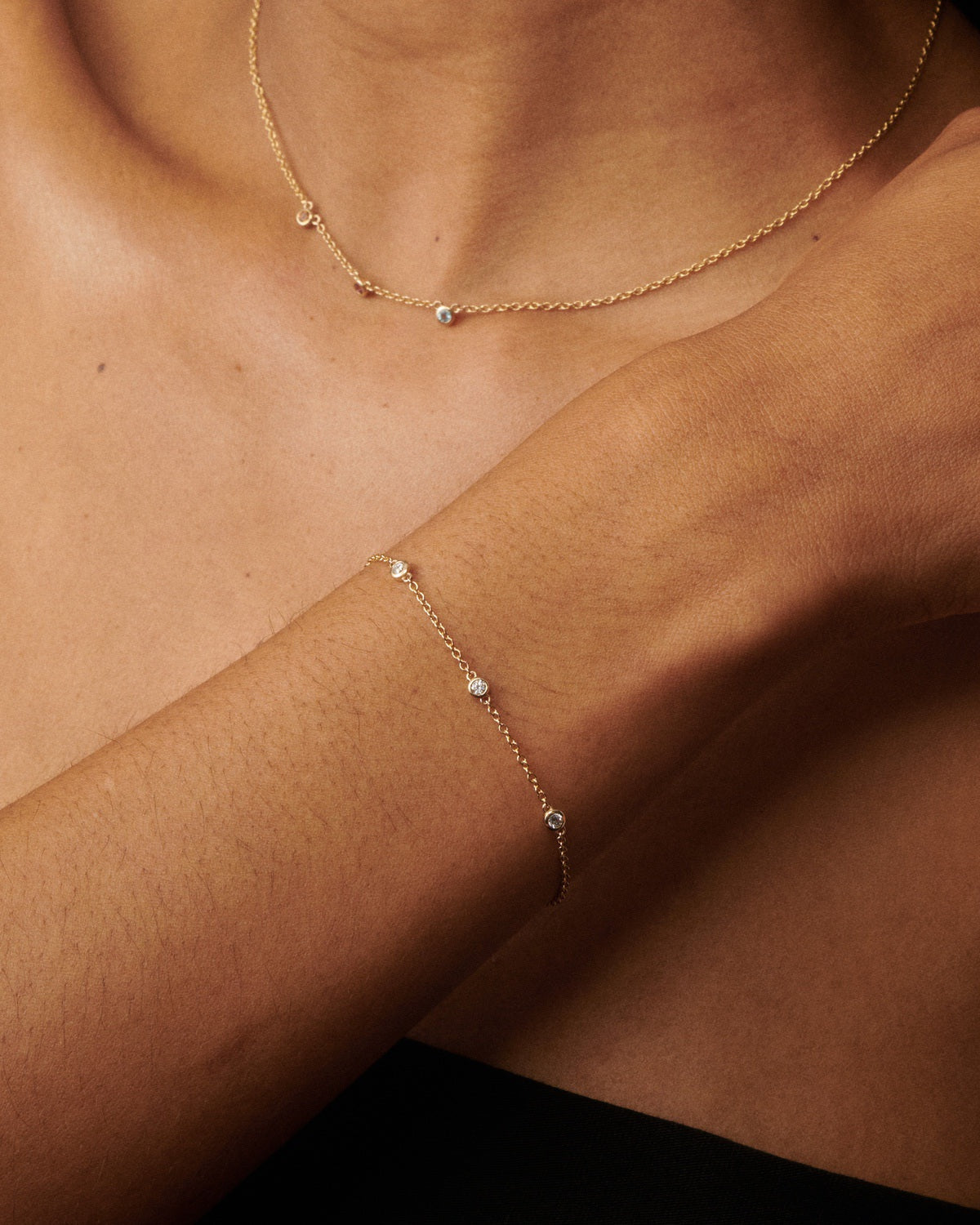 A close-up of a model's neck and wrist wearing a delicate gold chain necklace and a matching thin gold chain bracelet, each adorned with small clear gemstones.
