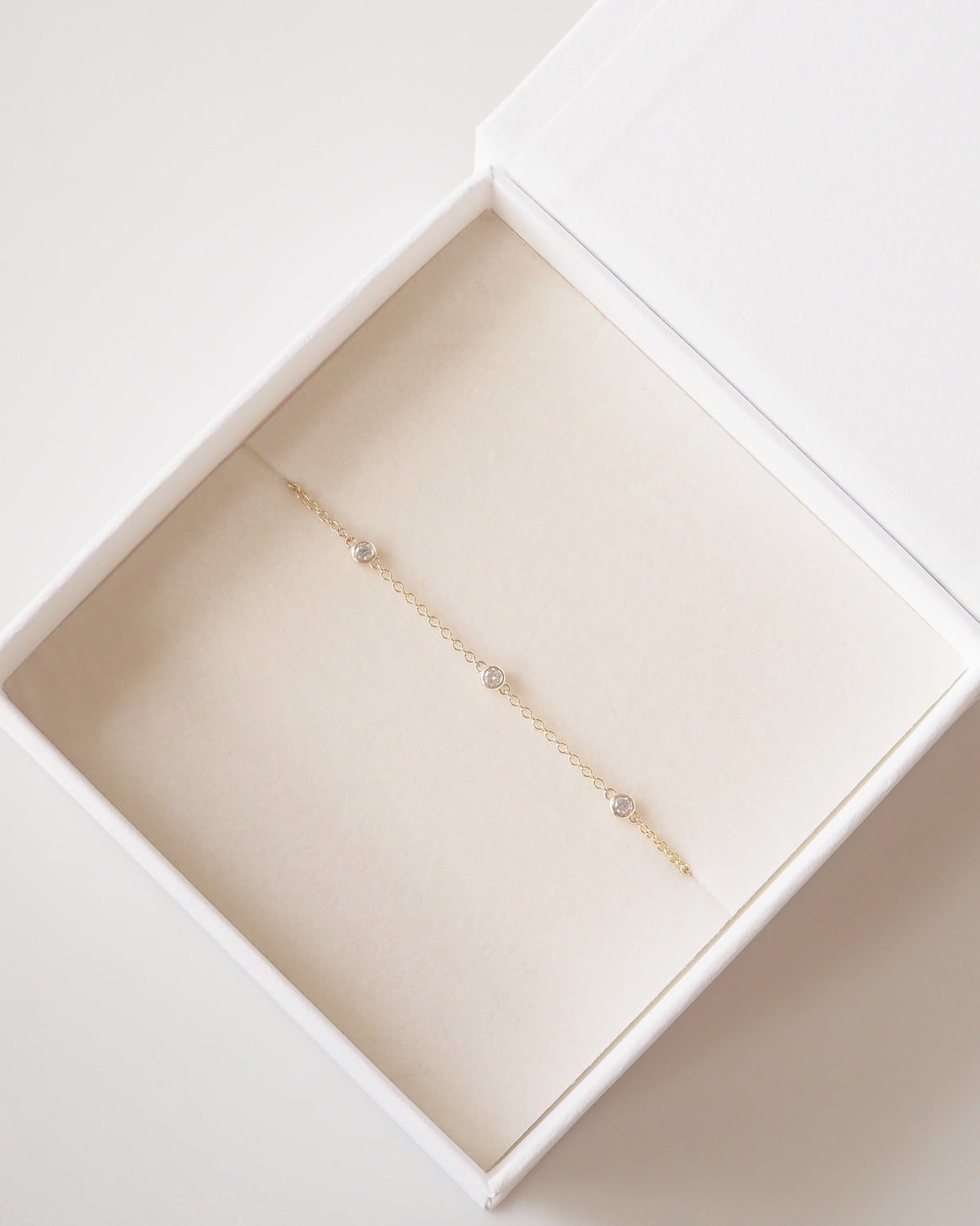 A delicate gold chain bracelet with three small clear gemstone accents, presented inside an open white jewelry box with a cream-colored velvet insert.