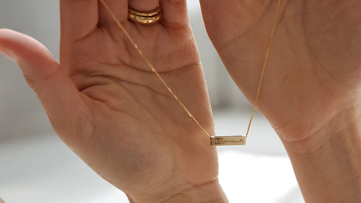 Gold necklace with a bar pendant held between two hands against a neutral background