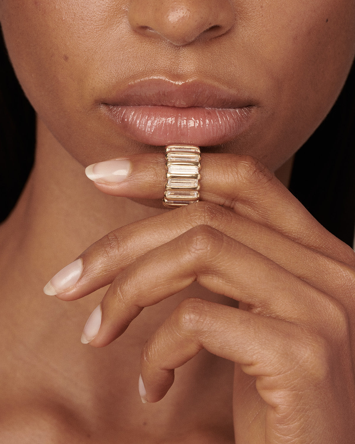 A close-up of a model's hand and lower face wearing a wide gold ring with rectangular baguette-cut stones, set against a dark background. 