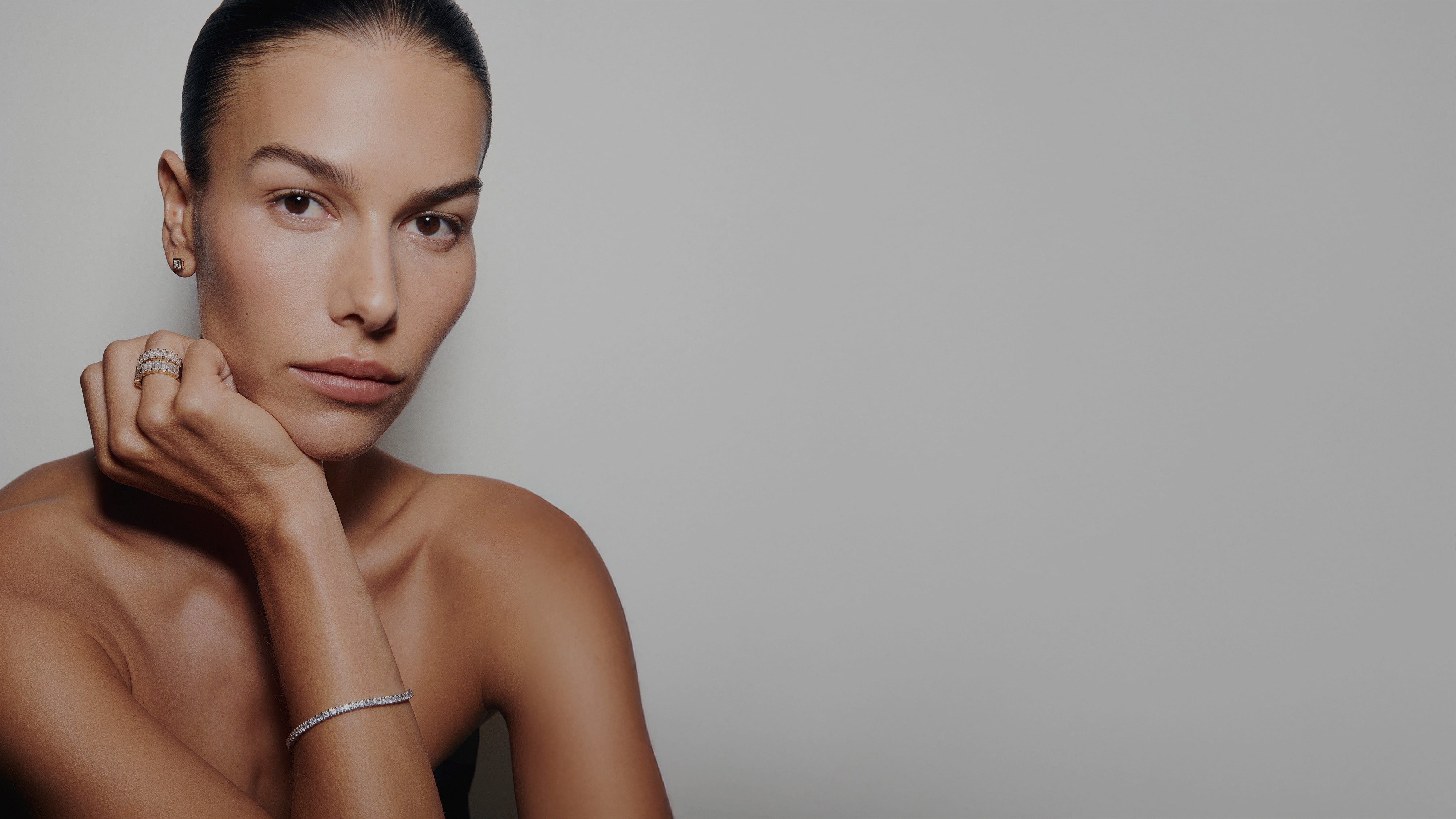 Close-up portrait of a woman wearing Kate & Kole lab-grown diamond pieces, including rings, stud earrings, and a delicate tennis bracelet, against a minimal neutral background.
