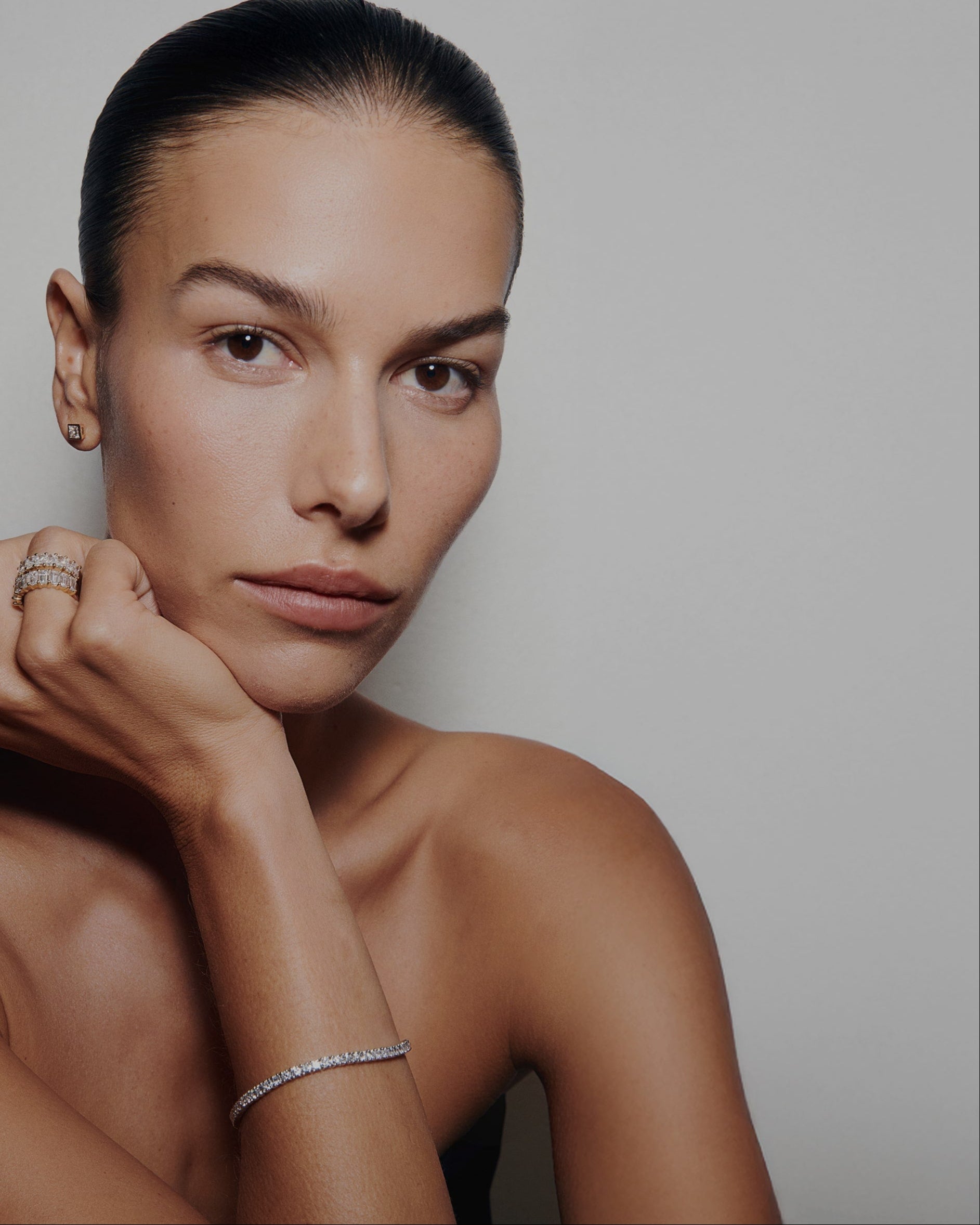 Close-up portrait of a woman wearing Kate & Kole lab-grown diamond pieces, including rings, stud earrings, and a delicate tennis bracelet, against a minimal neutral background.