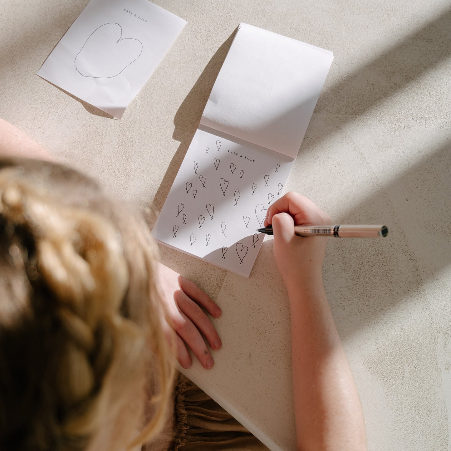 Person sketching heart designs in a notebook on a sunlit table, suggesting luxury jewellery conceptualisation.