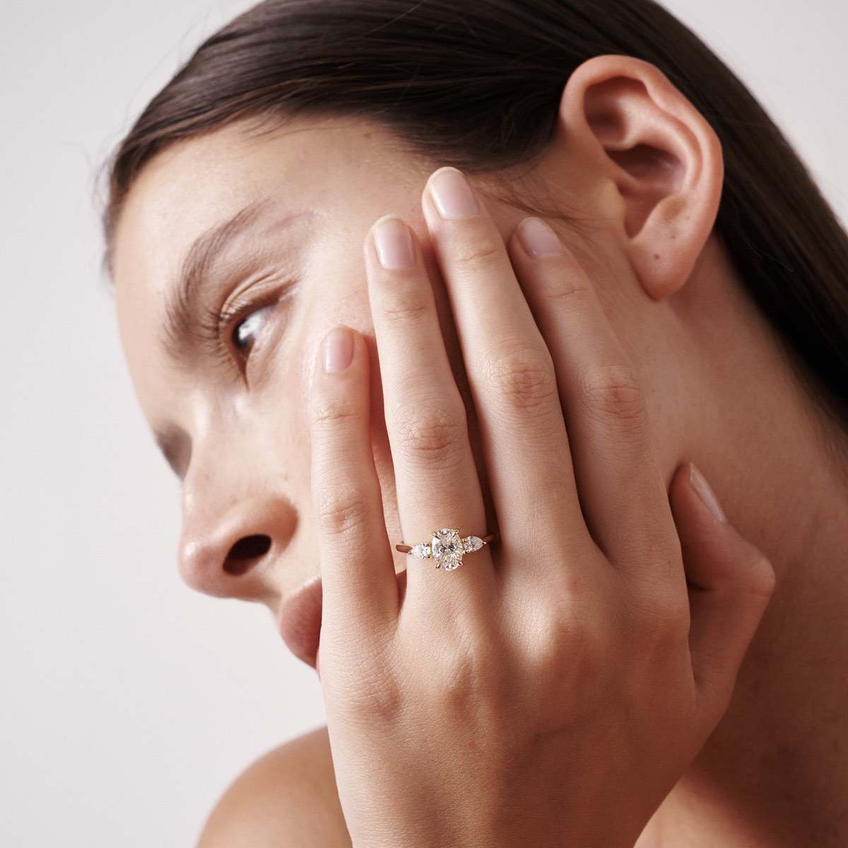 Our Ania ring showcases an oval centre stone shining between two smaller-sized pear shoulder stones. The ring pictured features a 1.00ct oval in 18ct yellow gold, sitting on the models hand and holding her face. 