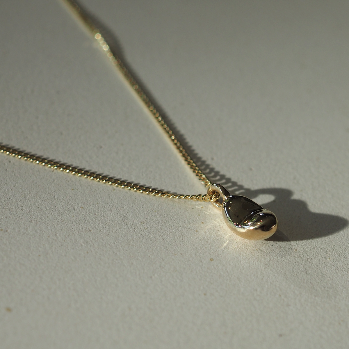 Elegant gold necklace with a smooth, teardrop pendant on a fine chain, casting a delicate shadow on a light surface.