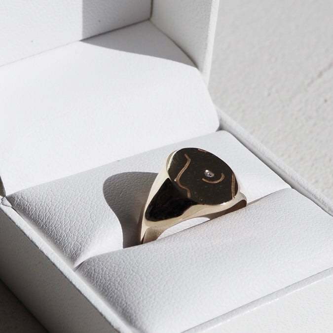 Gold and black signet ring with diamond accent in a white jewellery box, illuminated by sunlight. Luxurious and elegant design.