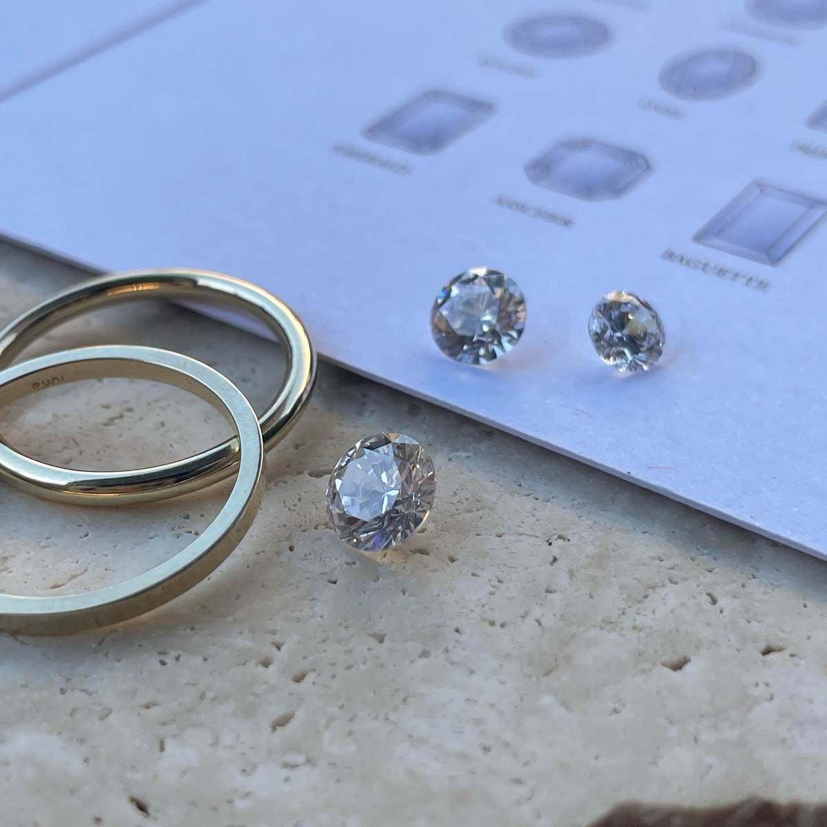 Gold rings and round-cut diamonds on a stone surface beside a gem guide. Luxury jewellery design elements.
