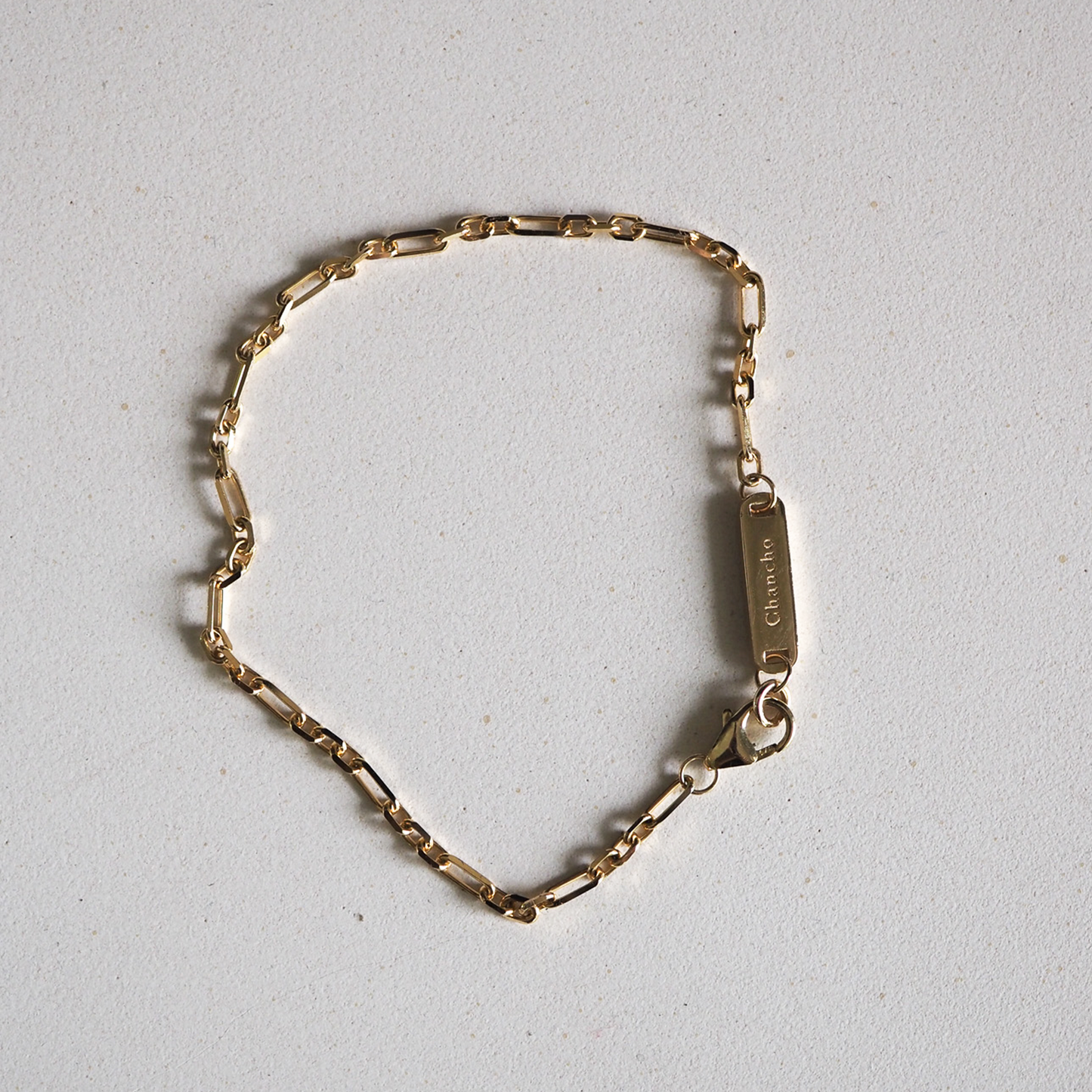 Gold chain bracelet with oval links and engraved rectangular tag on a light background, reflecting luxury designer jewellery.