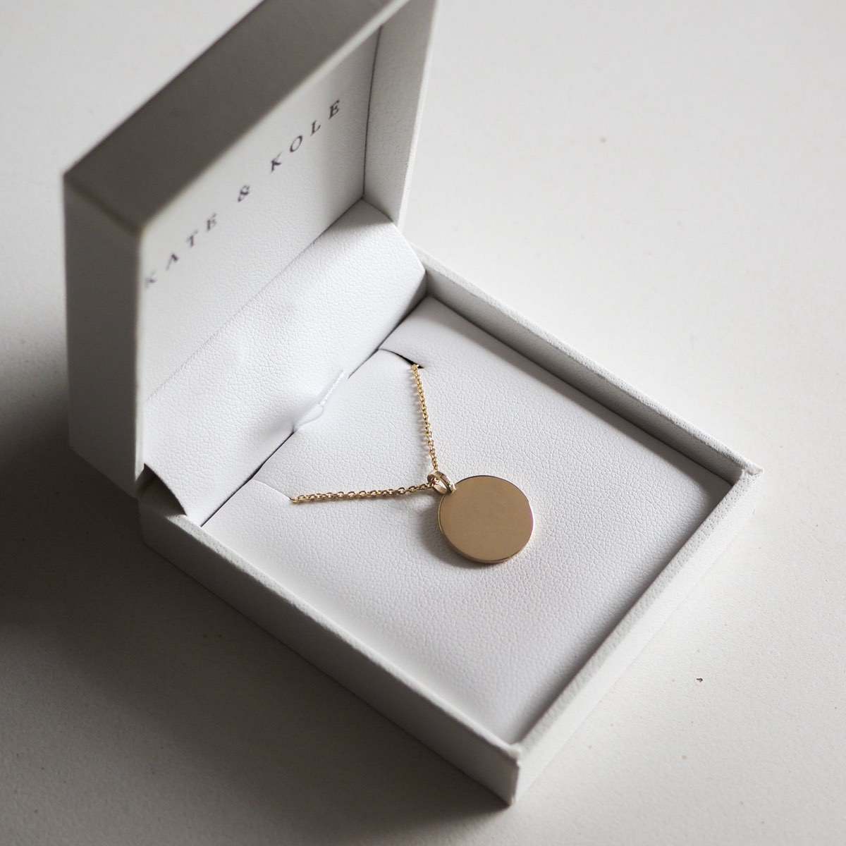 Gold pendant necklace in an open white jewellery box, featuring a round disc on a delicate chain. Luxury accessory in elegant packaging.
