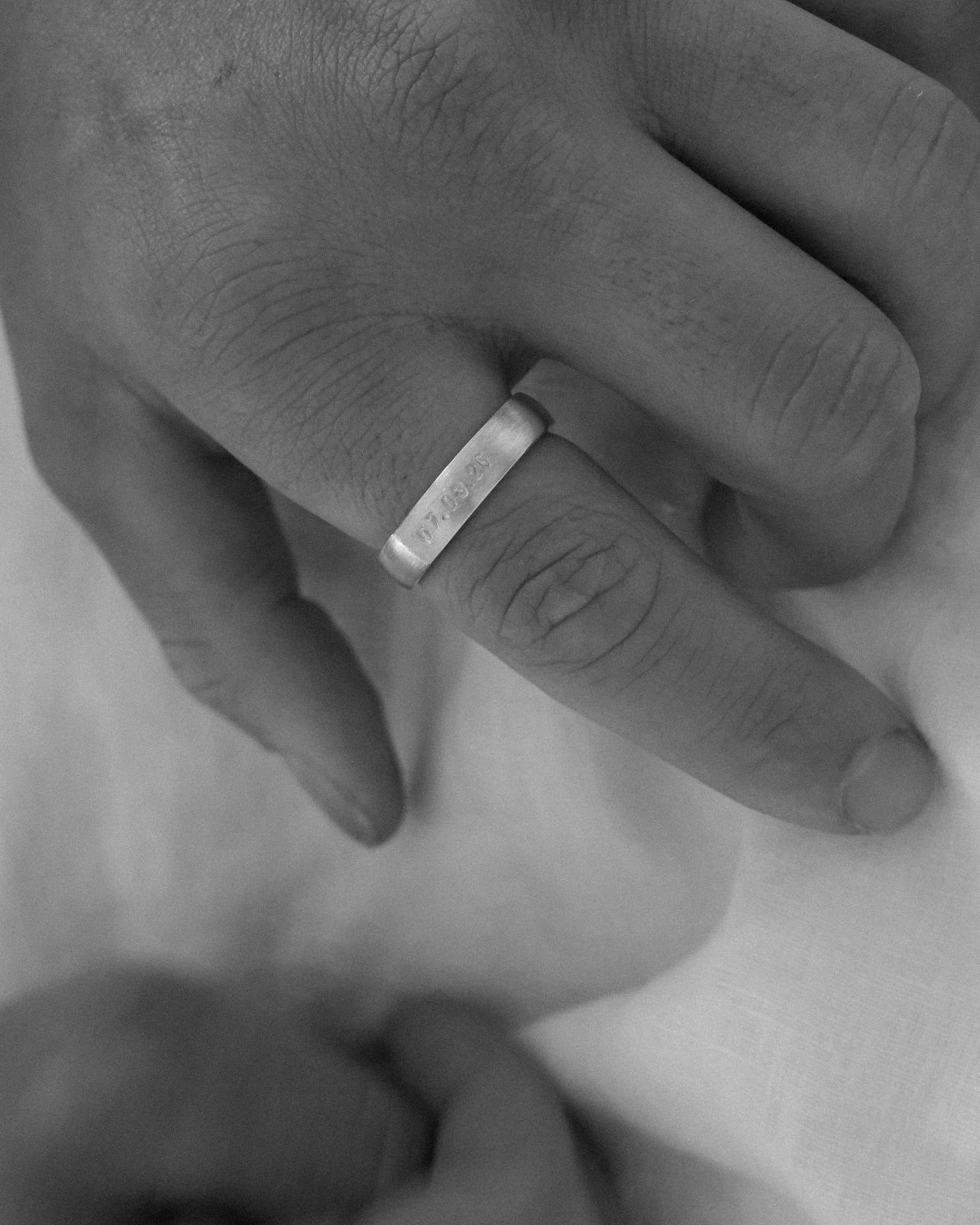 A close-up black and white photograph of a person's hand wearing a simple metal band ring on one finger, resting on a soft white fabric.