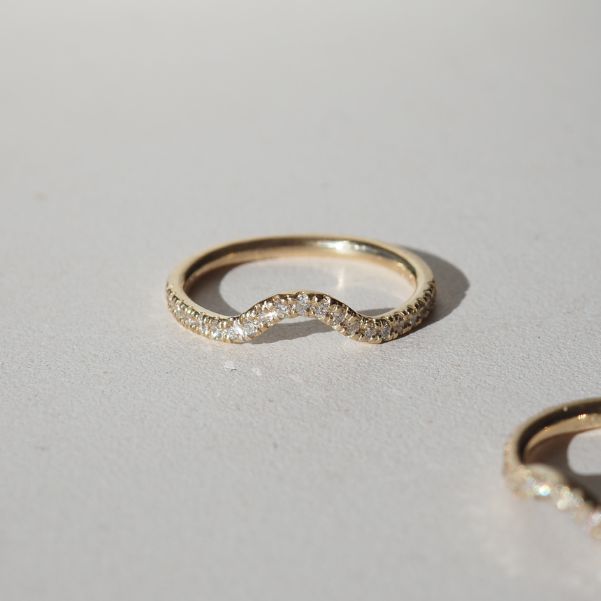 Gold ring with a subtle wave design, encrusted with small sparkling diamonds, photographed on a light surface. Luxury jewellery detail.