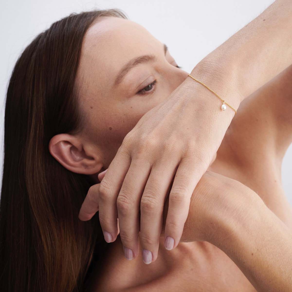 Woman with long brown hair wearing an elegant gold bracelet with a pearl, positioned gracefully near her face against a plain background.