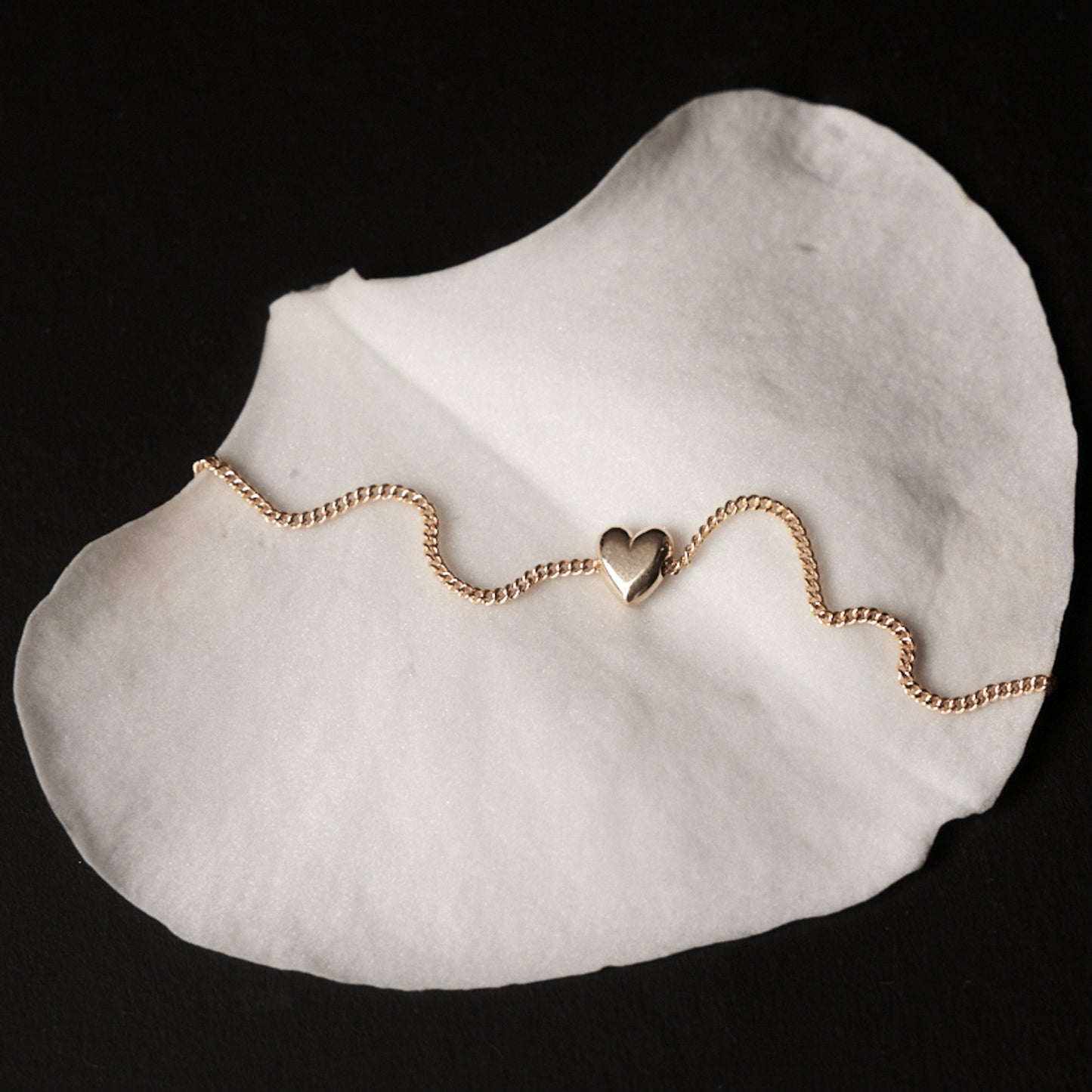 Gold bracelet with a heart charm on a white petal against a black background