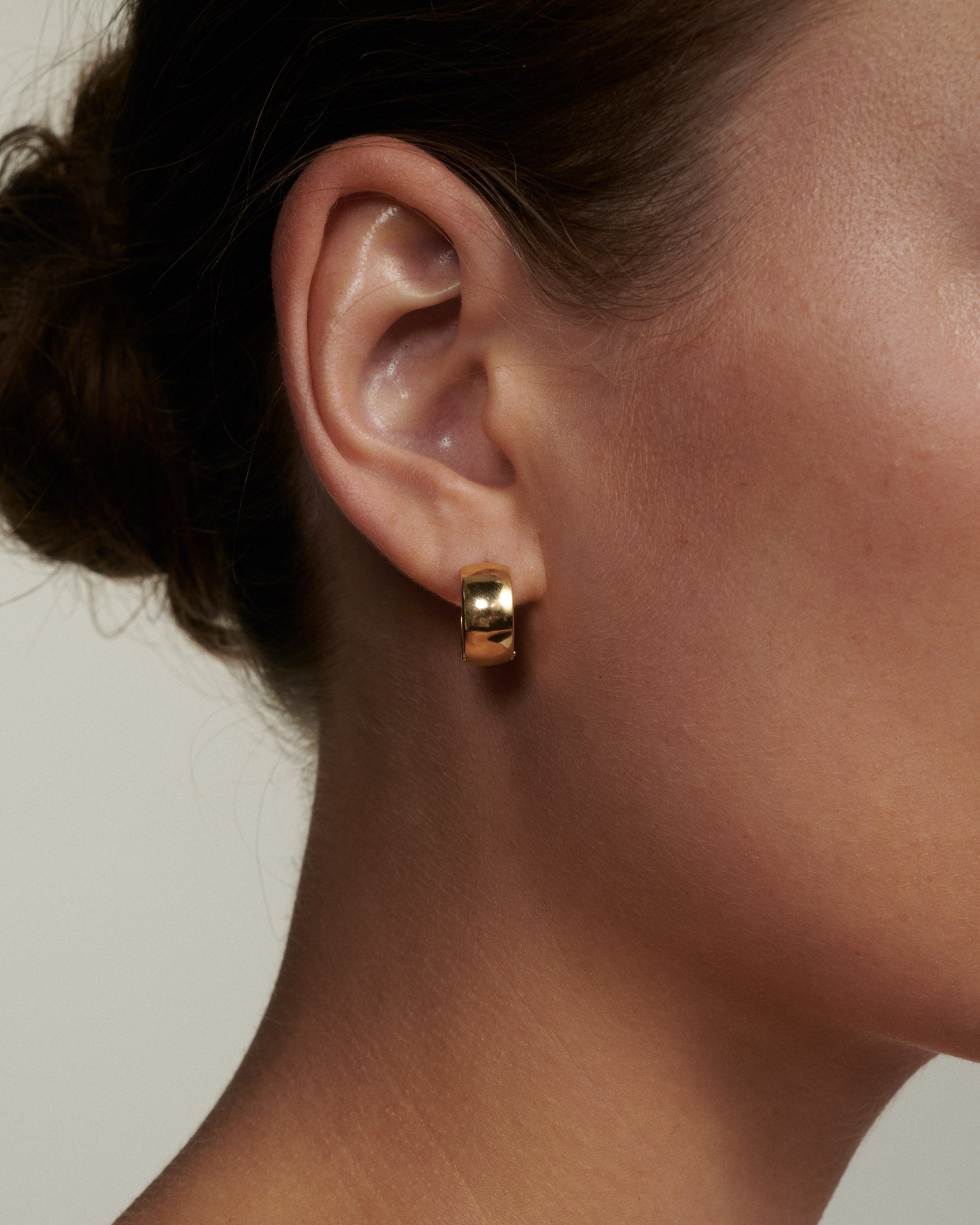 Close-up of an ear wearing a gold hoop earring against a neutral background