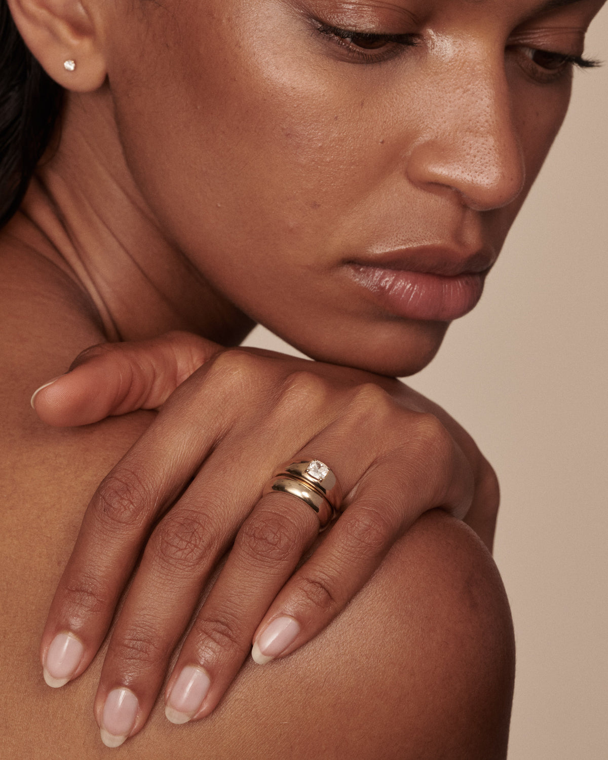 A close-up lifestyle shot of a woman wearing a stack of two gold rings, one with a small clear round gemstone, on her ring finger. She is looking down and resting her hand on her shoulder against a solid beige background.