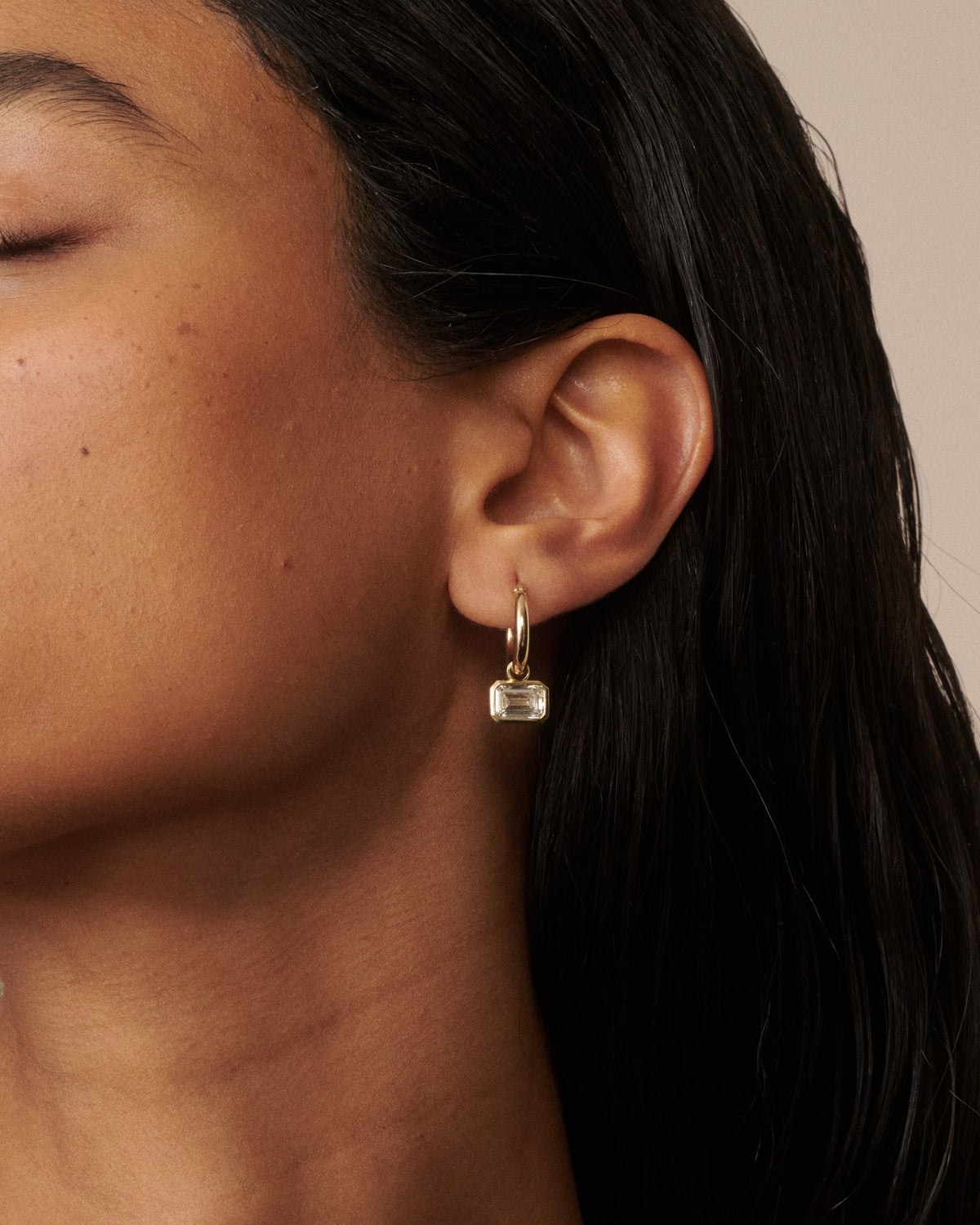 A close-up of a model's ear wearing a delicate gold huggie hoop earring with a small clear faceted rectangular drop stone, set against a warm beige background. [text overlay: Francielle wears 4ct total carat weight]