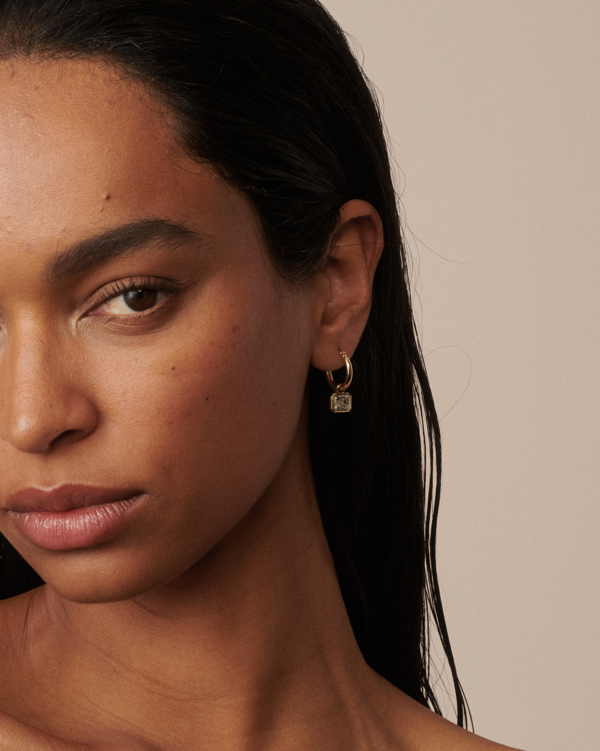 A close-up portrait of a woman with dark hair wearing small gold hoop earrings with clear diamond drops, set against a solid beige background. [text overlay: Francielle wears 4ct total carat weight]