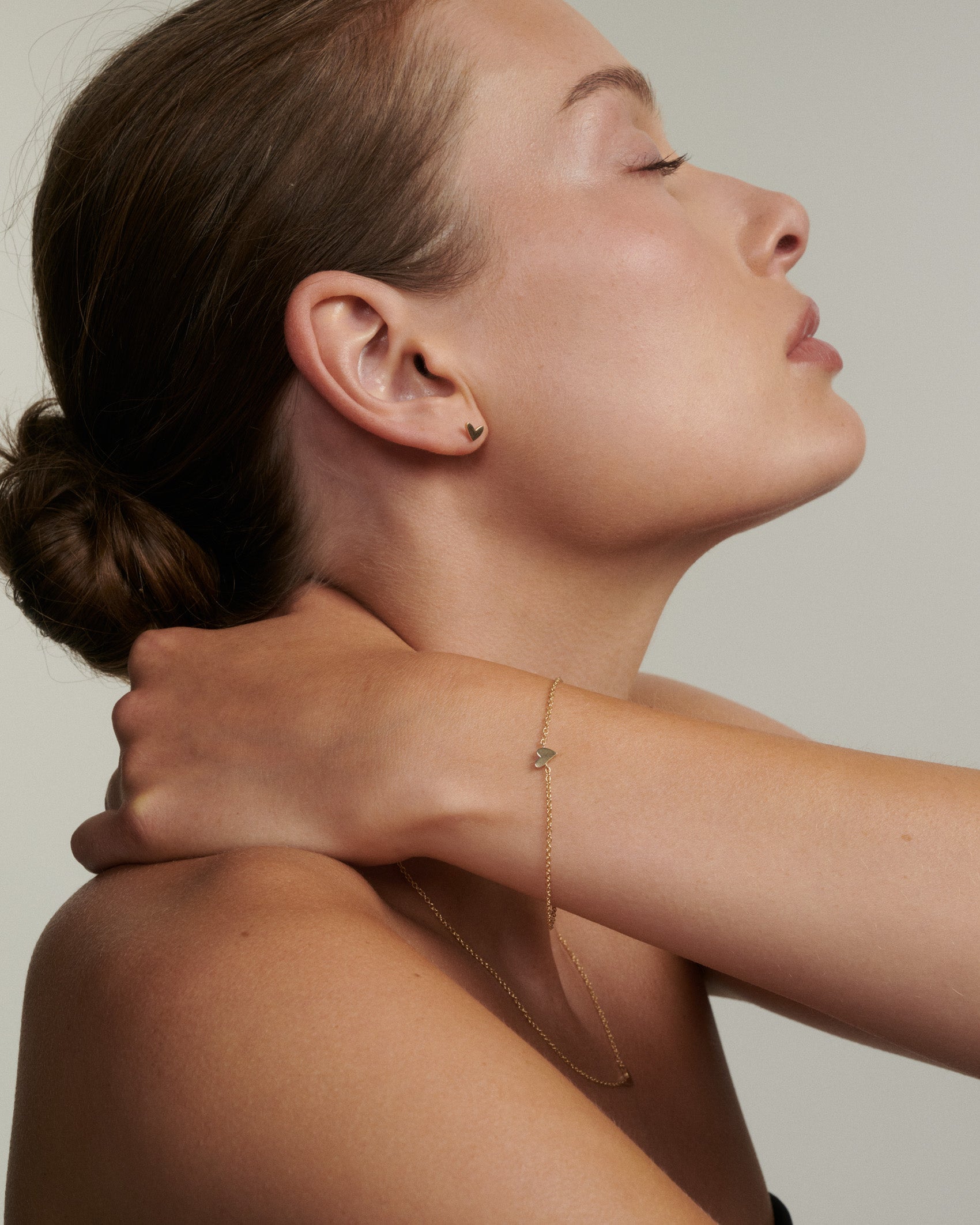Woman wearing a delicate gold necklace against a neutral background [text overlay: Andrea wears the Hand Drawn Heart Bracelet, Studs & Necklace]