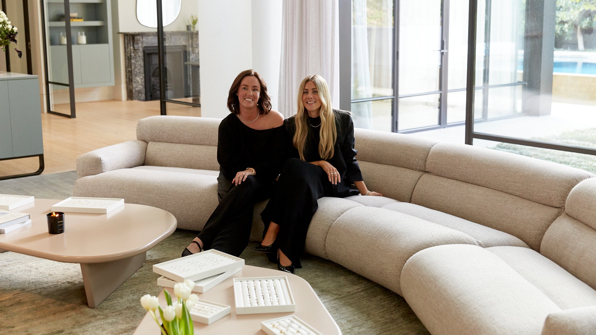 Two women in black outfits sit on a curved beige sofa in a luxurious, sunlit living room. Open jewellery boxes are on the coffee table.