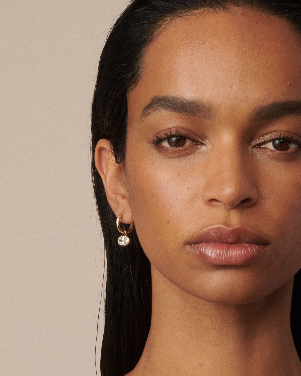 A close-up portrait of a model wearing a delicate gold hoop earring with a small clear diamond pendant against a solid beige background. [text overlay: Francielle wears 4ct total carat weight]