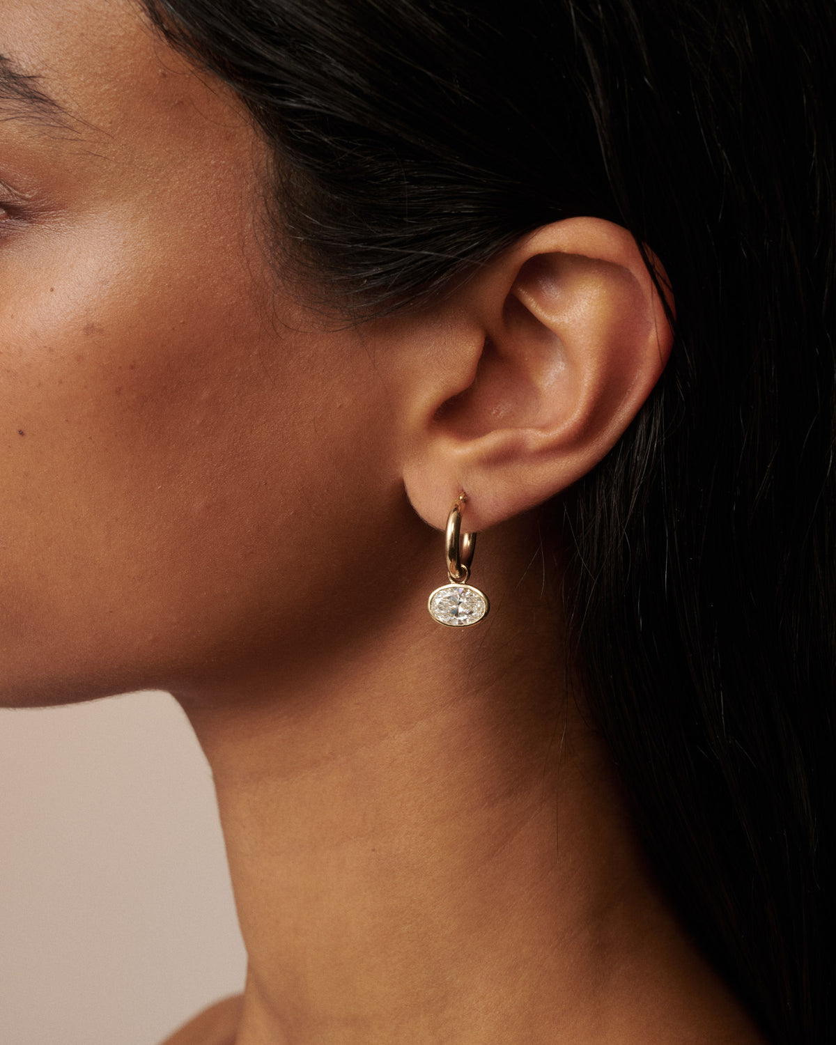 A close-up portrait of a woman with dark hair wearing small gold hoop earrings with clear diamond drops, set against a solid beige background. [text overlay: Francielle wears 4ct total carat weight]