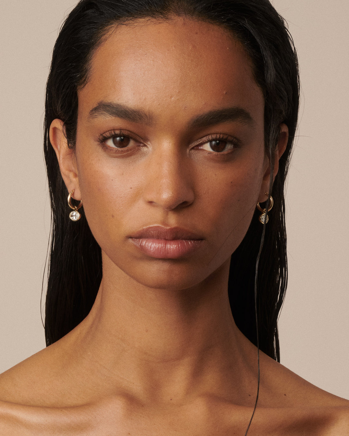 A close-up portrait of a woman with dark hair wearing small gold hoop earrings with clear diamond drops, set against a solid beige background. [text overlay: Francielle wears 4ct total carat weight]