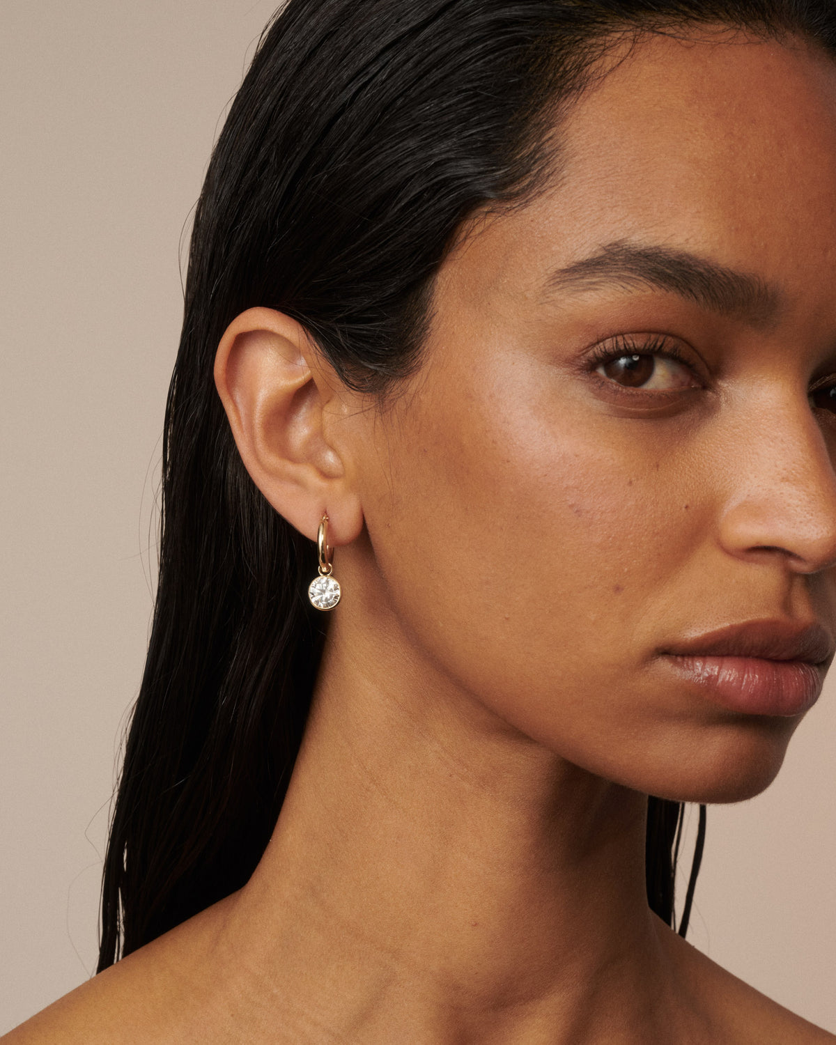 A close-up of a model wearing a delicate gold hoop earring with a small clear faceted gemstone dangle against a warm beige background. [text overlay: Francielle wears 4ct total carat weight]