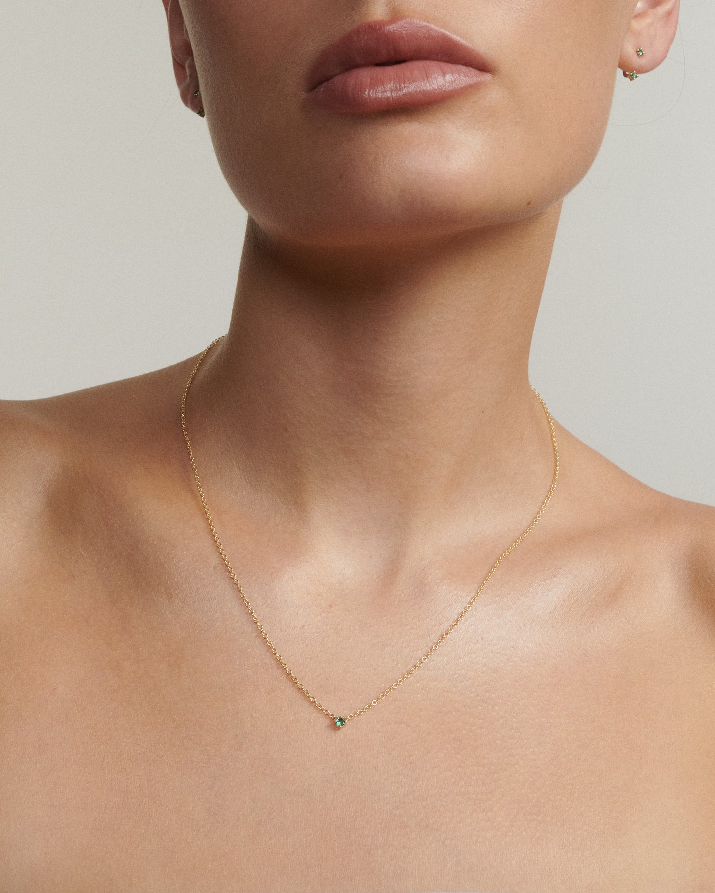 Close-up of a person wearing a delicate gold necklace with a small green gemstone on a neutral background.