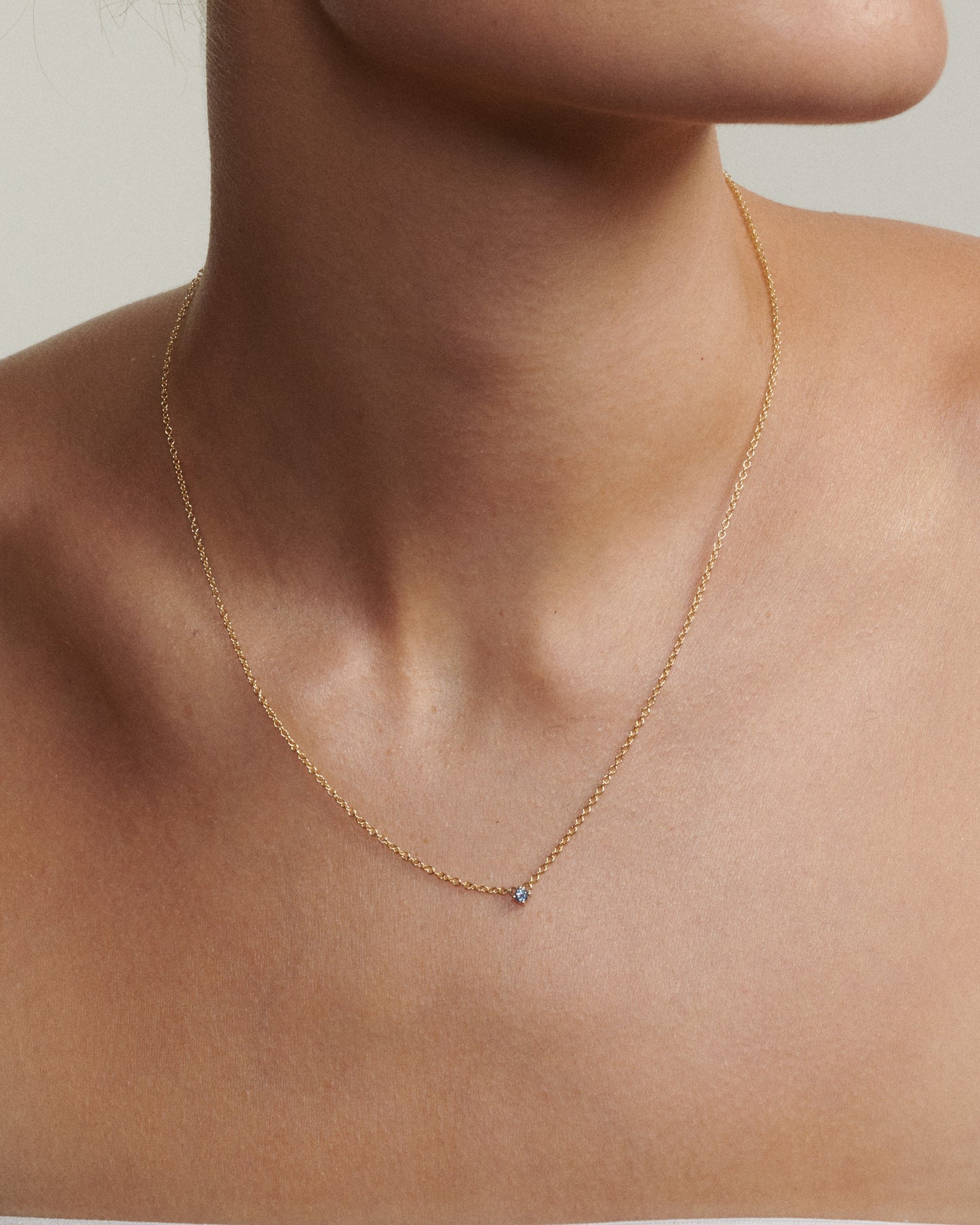 Gold necklace with a small blue gemstone on a person's neck against a neutral background