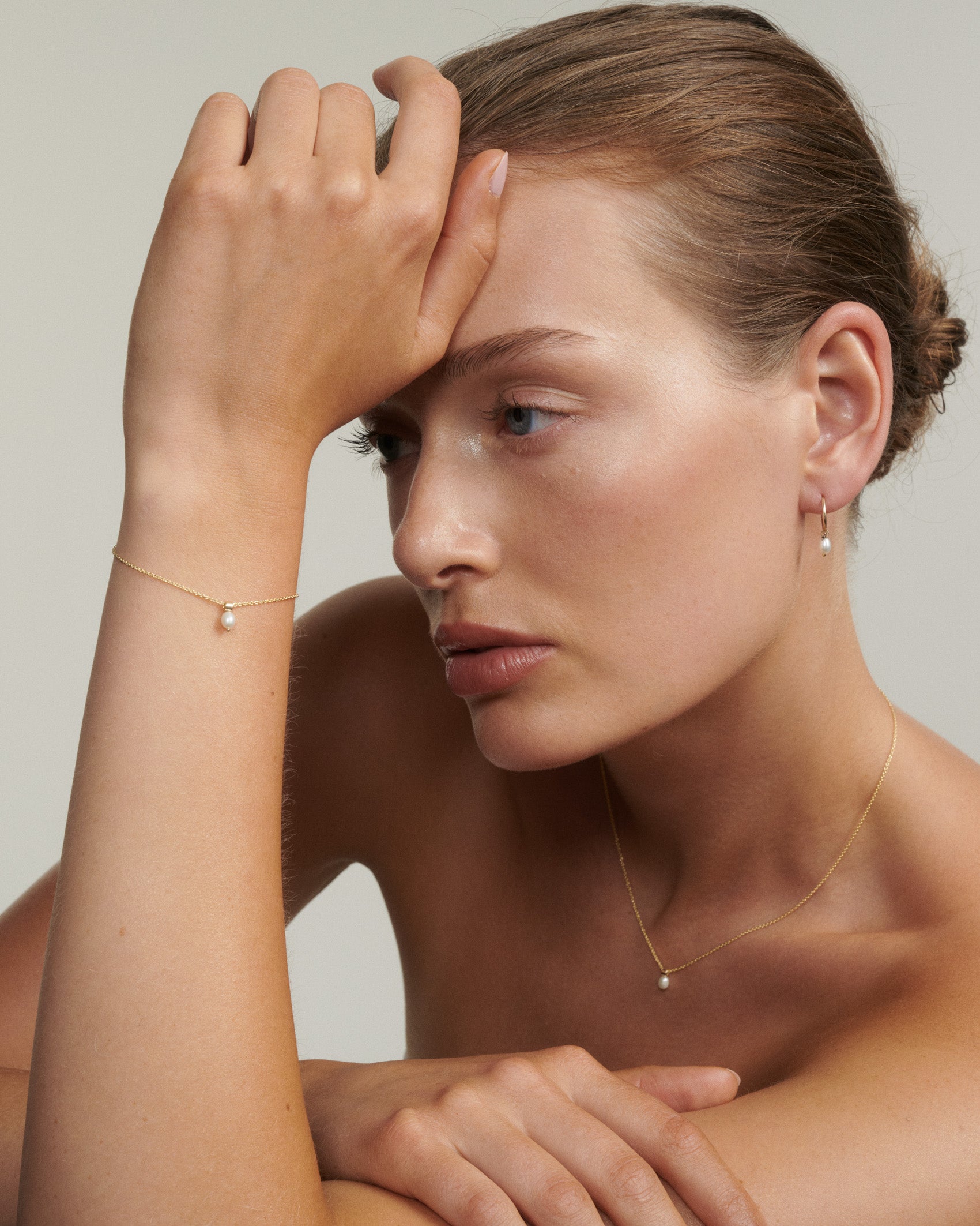 Woman wearing gold jewelry with a plain background [text overlay: Andrea wears the Tiny Pearl Bracelet, Necklace & Hoops]