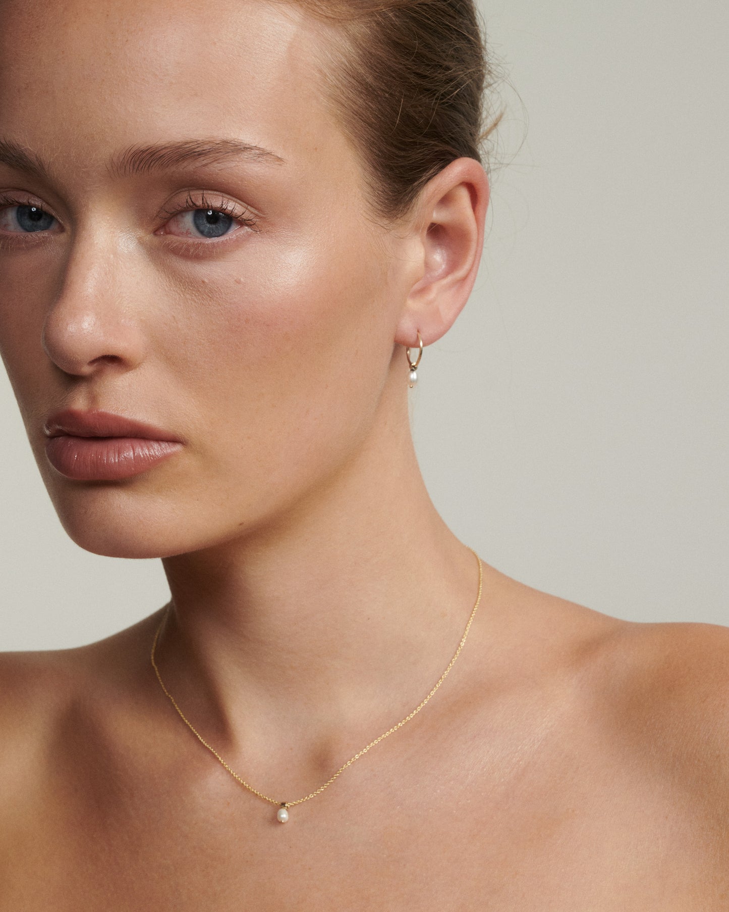 Woman wearing a gold necklace and earring on a neutral background [text overlay: Andrea wears the Tiny Pearl Studs & Necklace]