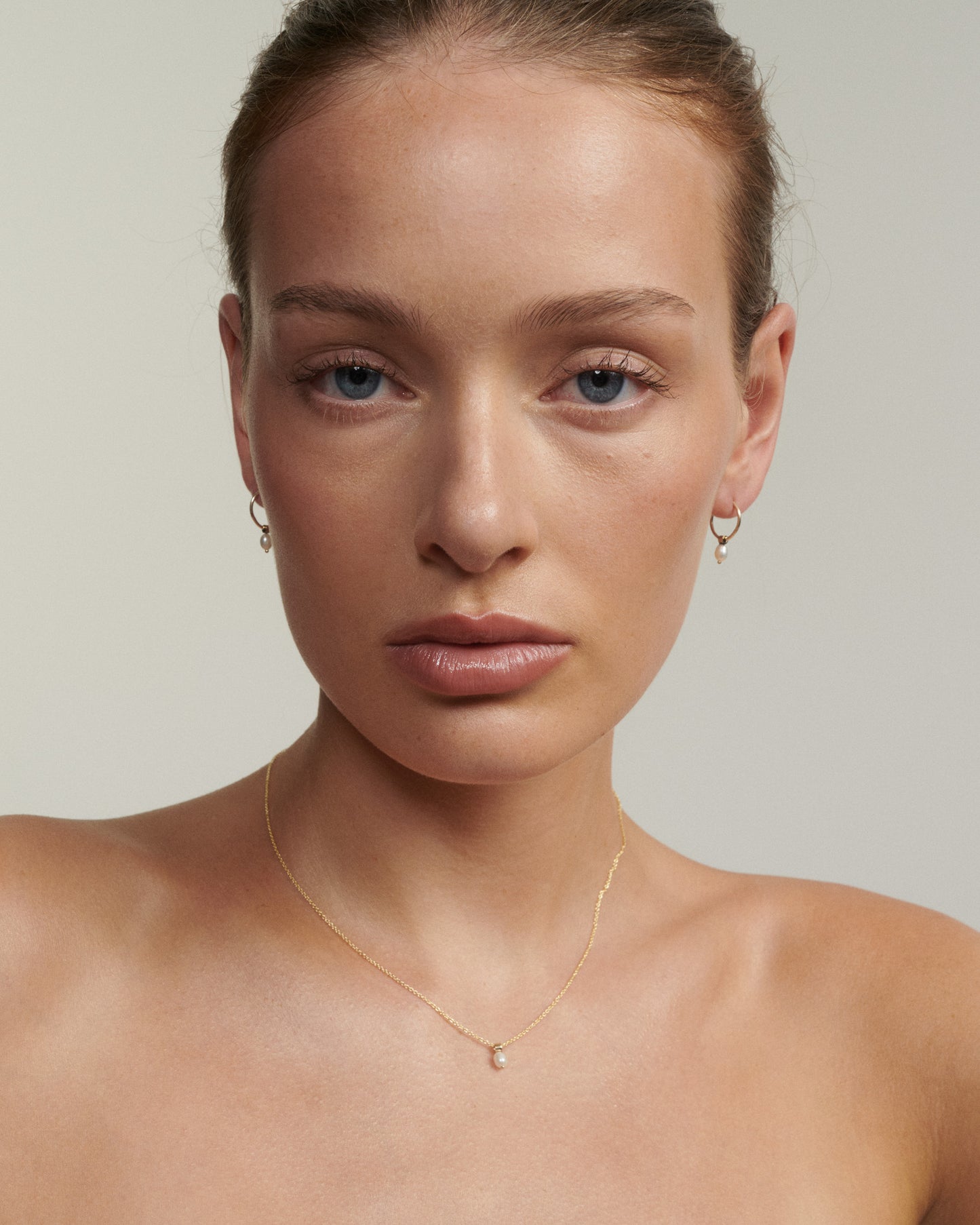 Woman wearing a gold necklace and earrings against a neutral background [text overlay: Andrea wears the Tiny Pearl Studs & Necklace]