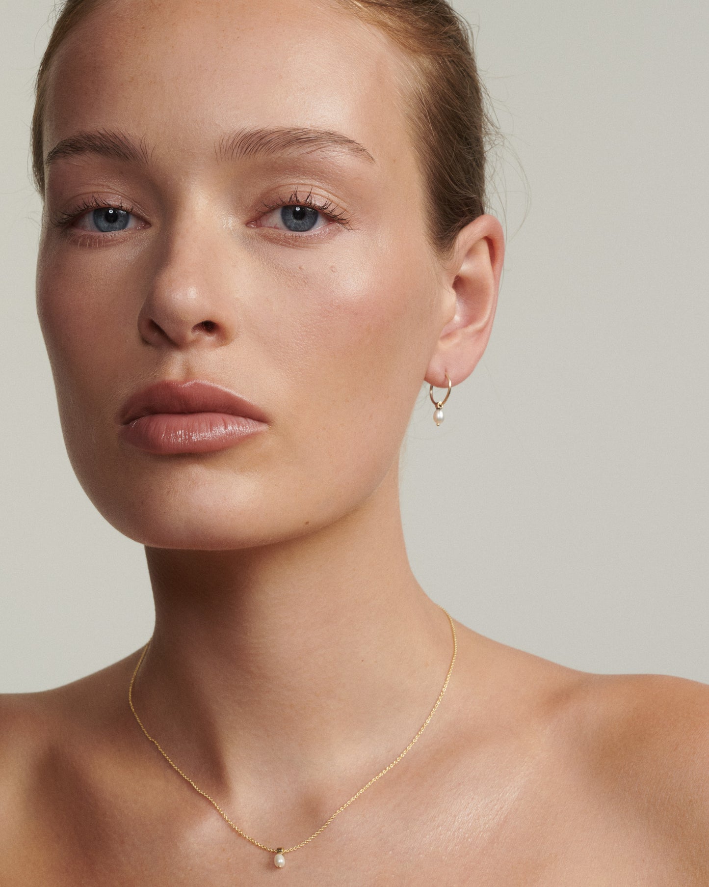 Close-up of a person wearing a gold necklace and earring on a neutral background
