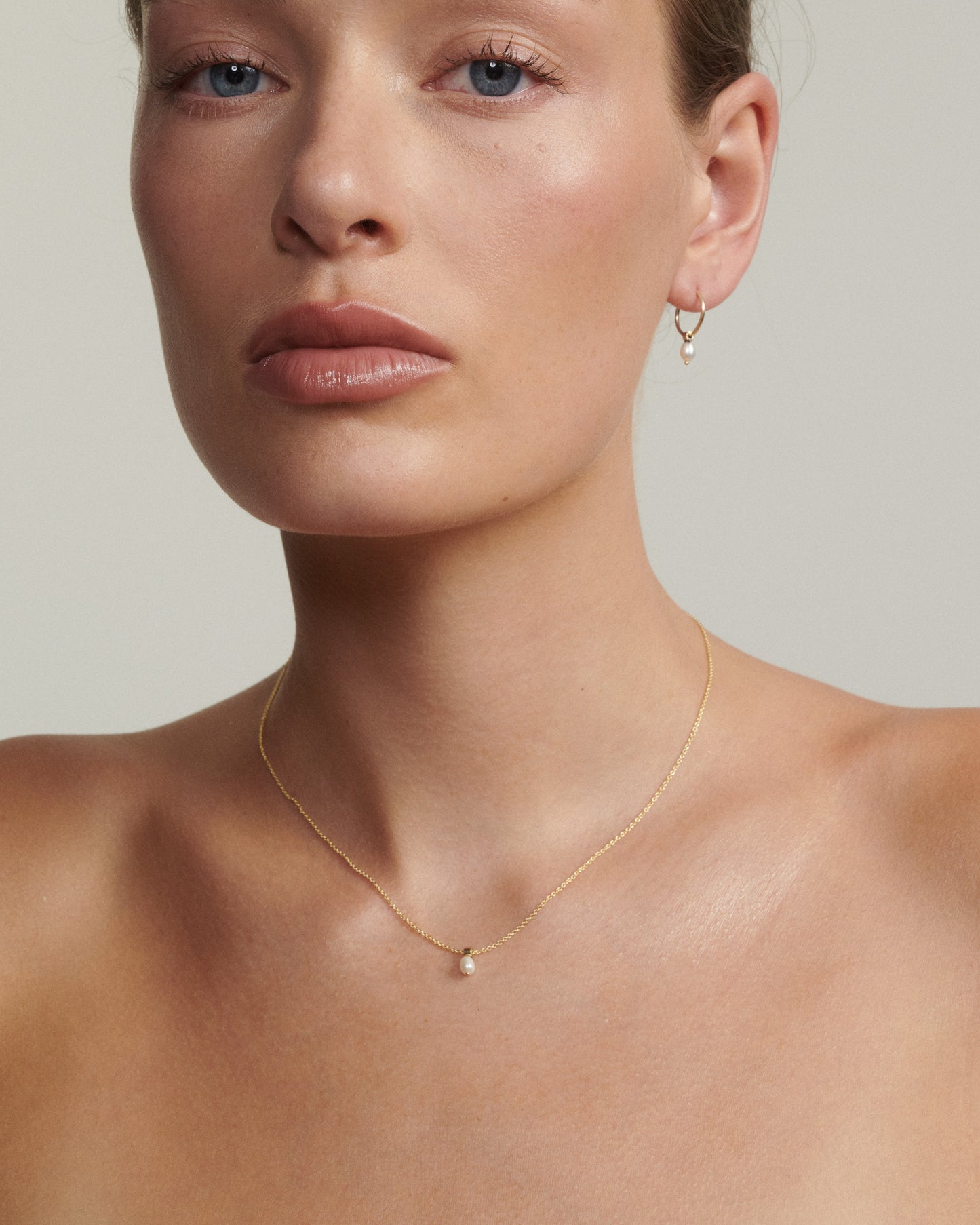 Close-up of a woman wearing a gold necklace and earring on a neutral background [text overlay: Andrea wears the Tiny Pearl Necklace & Hoops]