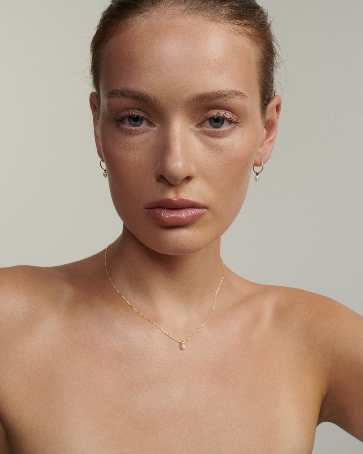 Woman wearing a gold necklace and earrings against a neutral background [text overlay: Andrea wears the Tiny Pearl Necklace & Hoops]