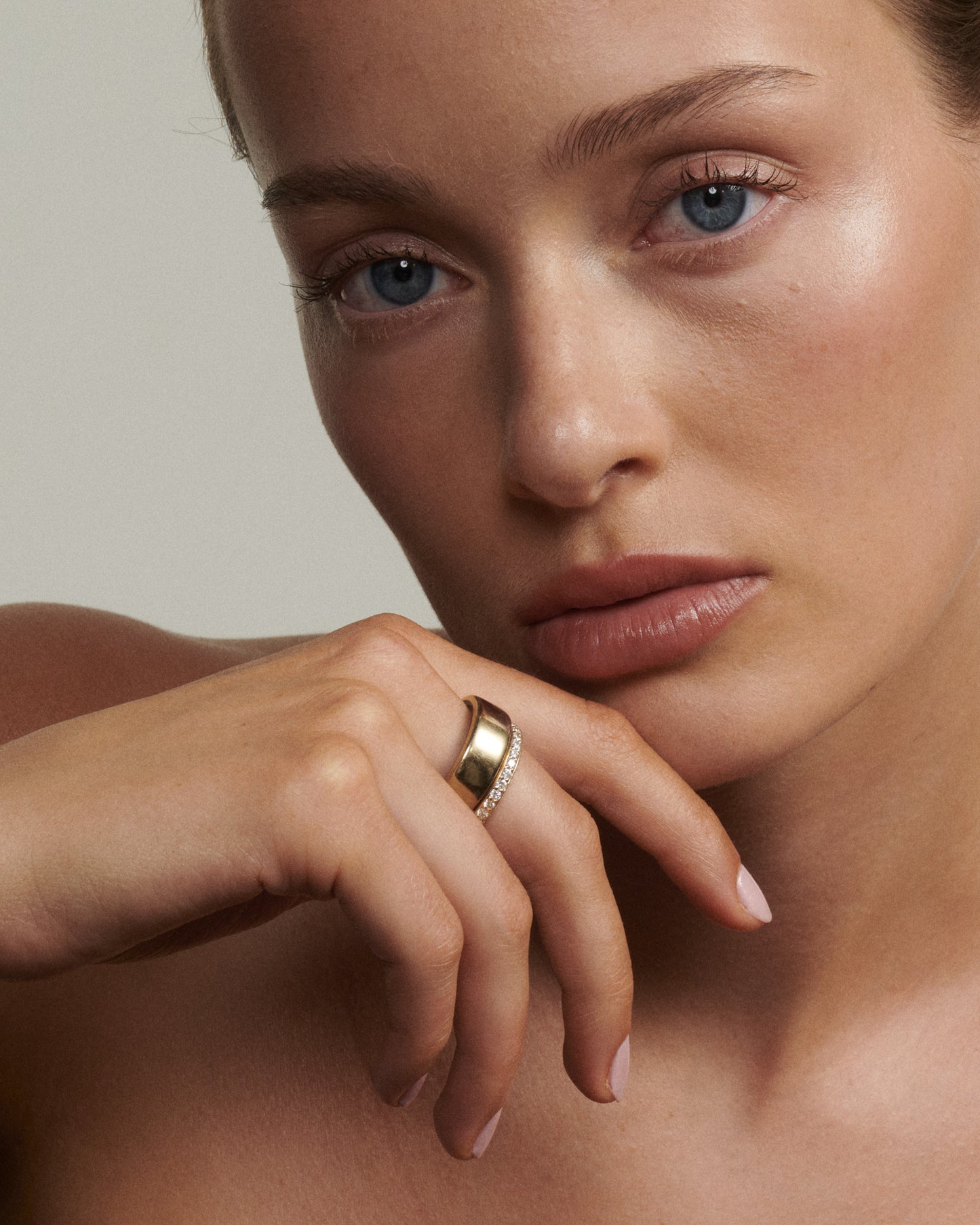 Close-up of a woman's face with a gold ring on her finger against a neutral background