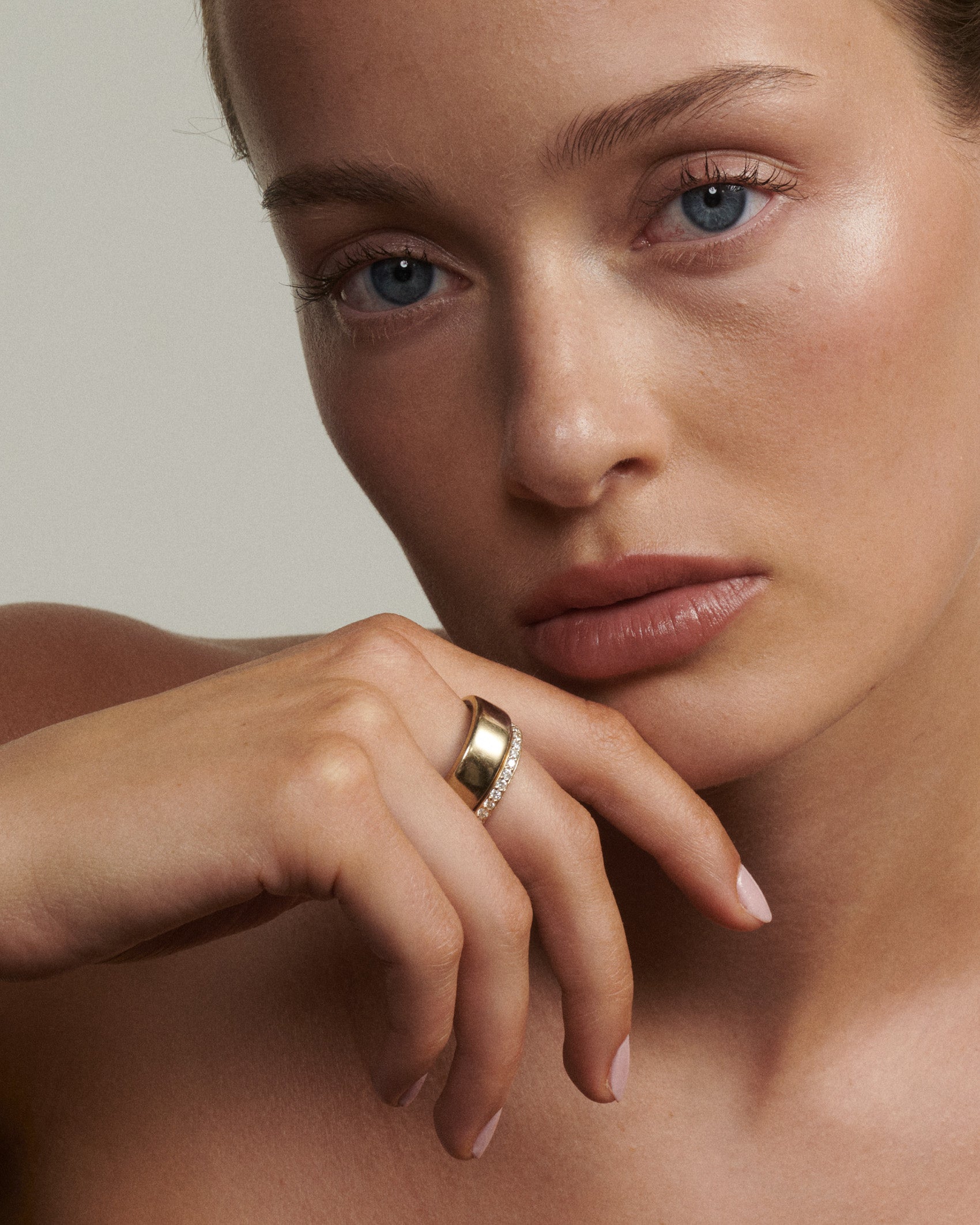Close-up of a woman's face with a gold ring on her finger against a neutral background  [text overlay: Andrea wears the Half Set Wellness Ring (9ct Yellow Gold, 7) stacked with the Oura Ring]