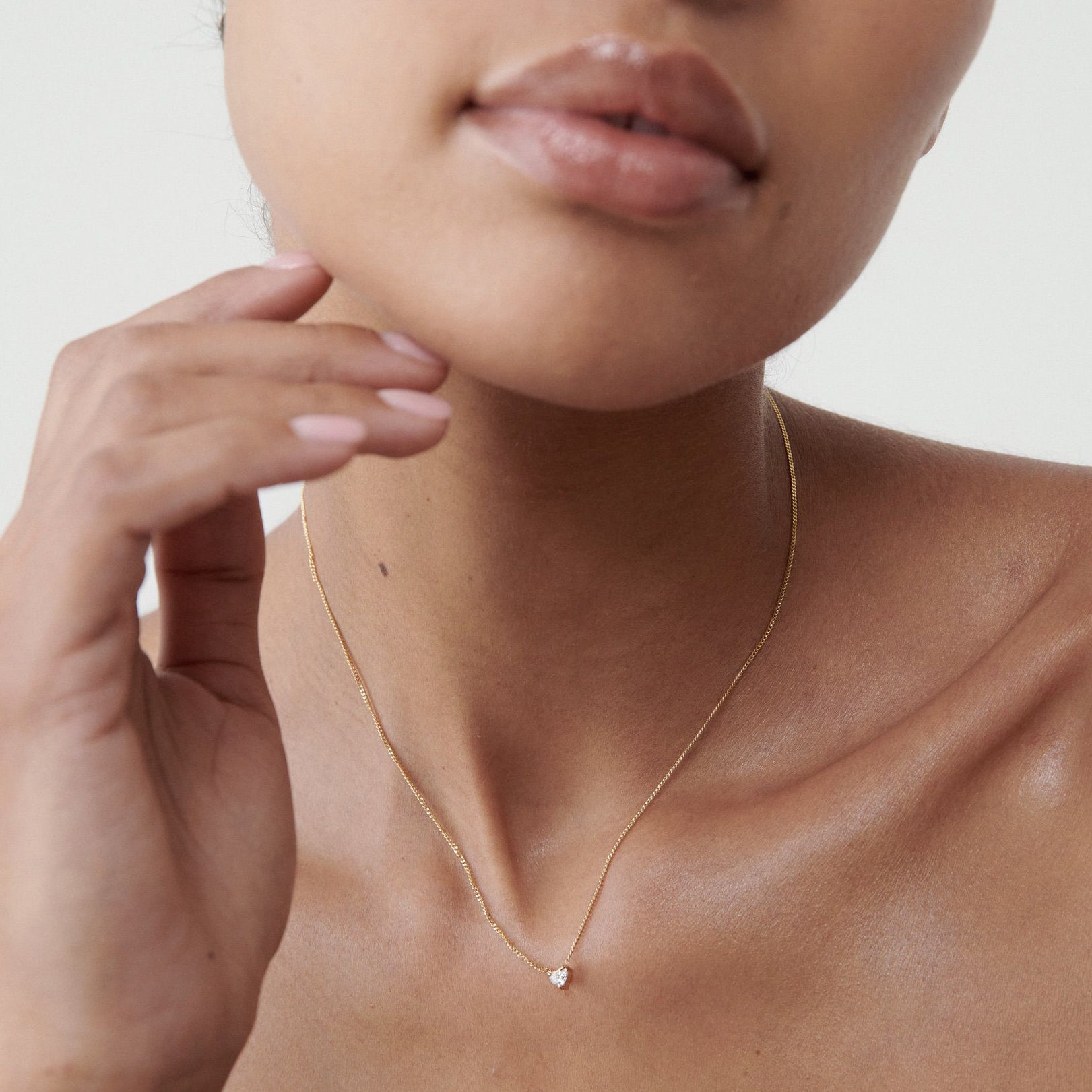 Woman wearing a delicate gold necklace with a small diamond pendant, softly touching her chin; luxury jewellery close-up.