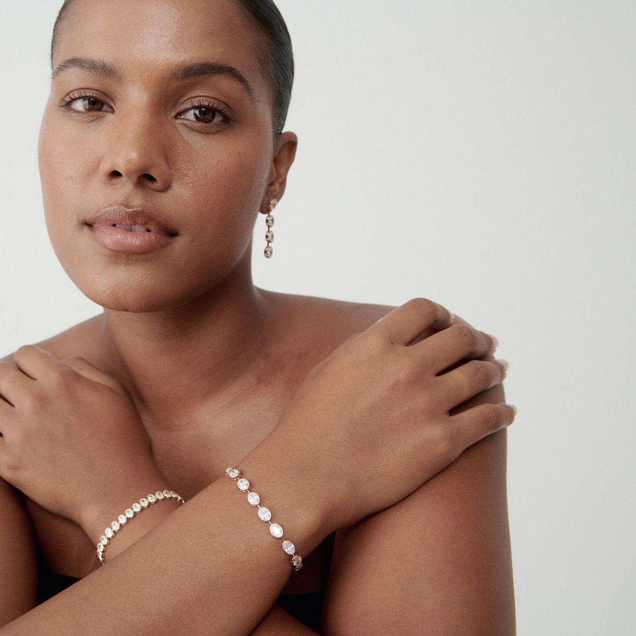 Woman wearing an elegant pearl bracelet and matching earrings, arms crossed, against a plain background, showcasing high-end jewellery.