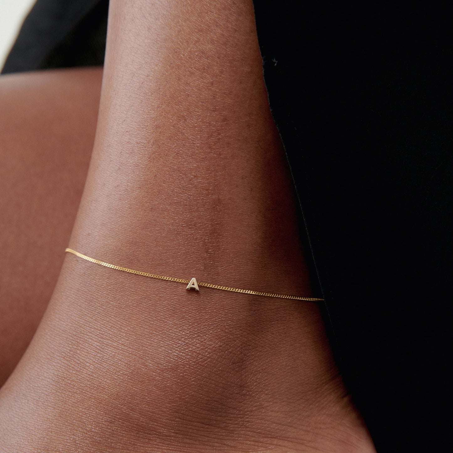 Close-up of a delicate gold ankle bracelet featuring a small letter 'A' charm on a person’s ankle, highlighting minimalist luxury jewellery.