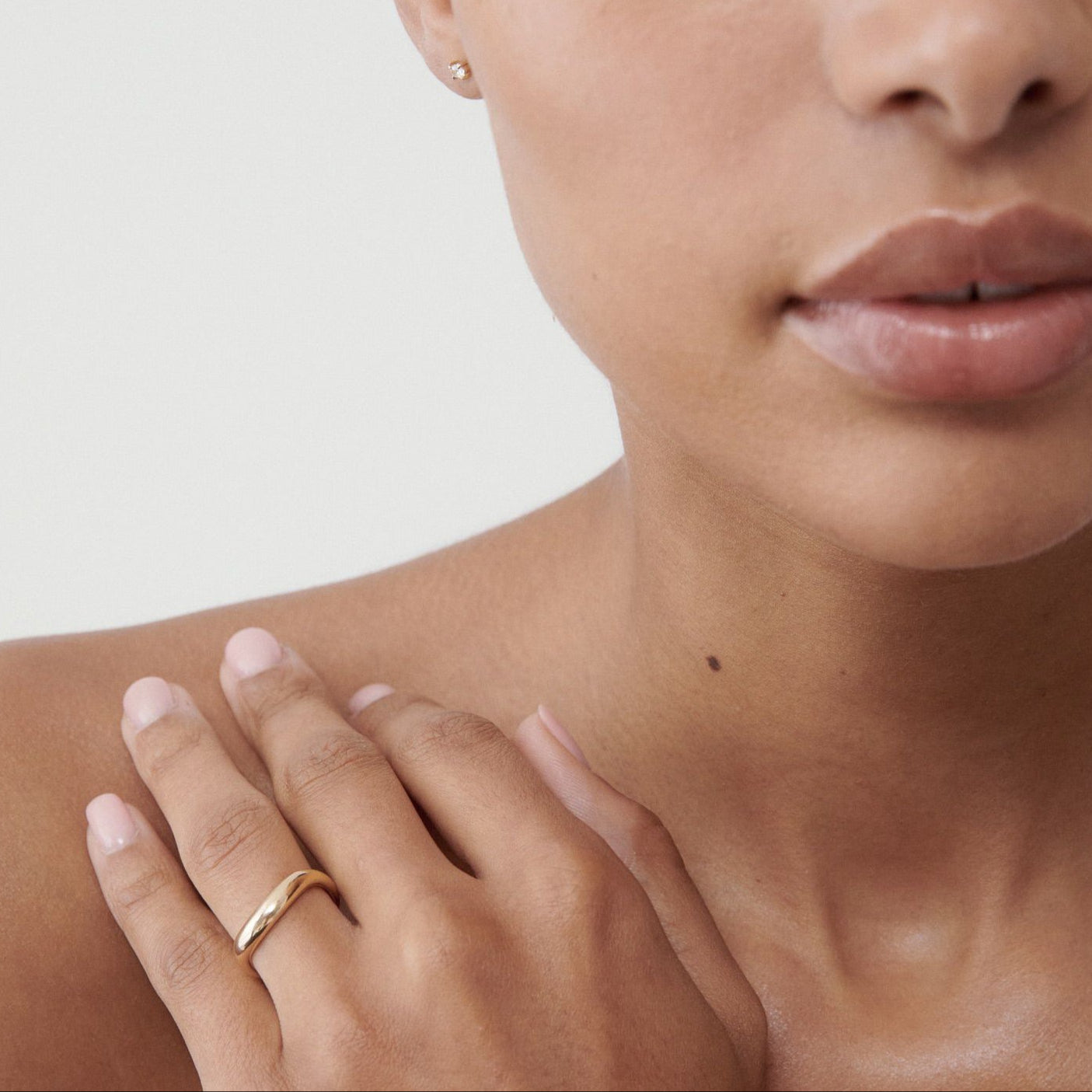 Close-up of a woman with gold earrings and a gold ring, showcasing luxury jewellery against a neutral background.