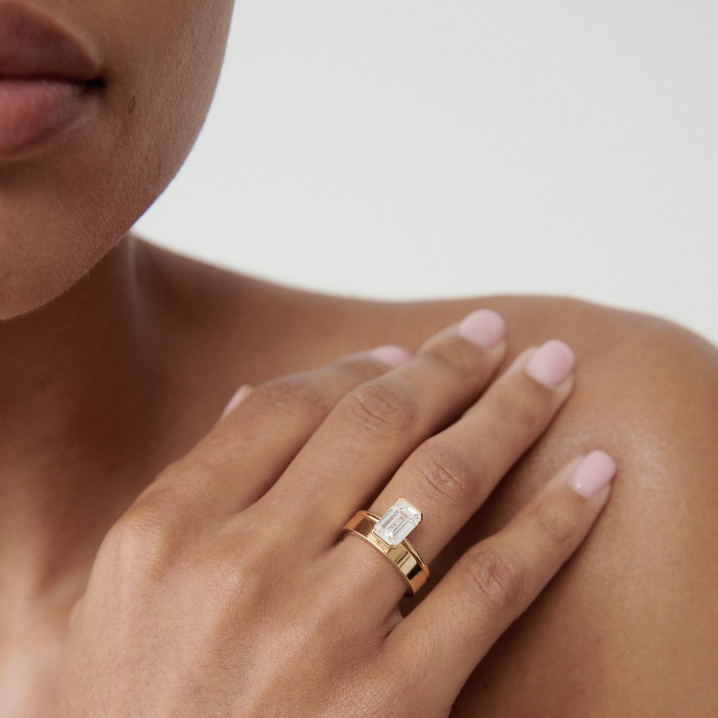 Close-up of a woman’s hand showcasing an emerald-cut diamond ring on a gold band, elegantly displayed against her shoulder.