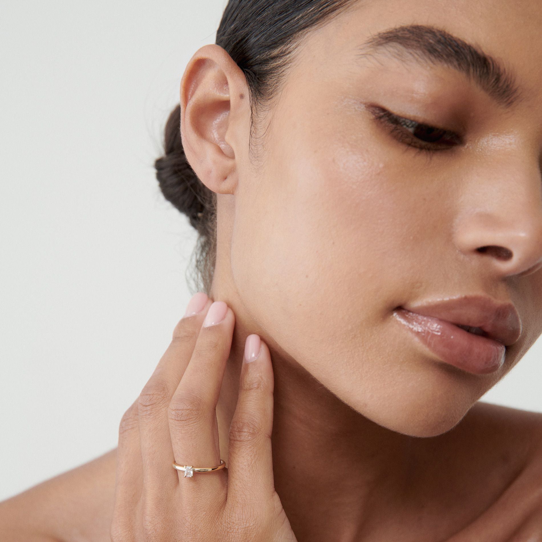Woman wearing an elegant gold ring with a small diamond, touching her face. Her hair is styled in a neat bun, showcasing minimalist luxury.