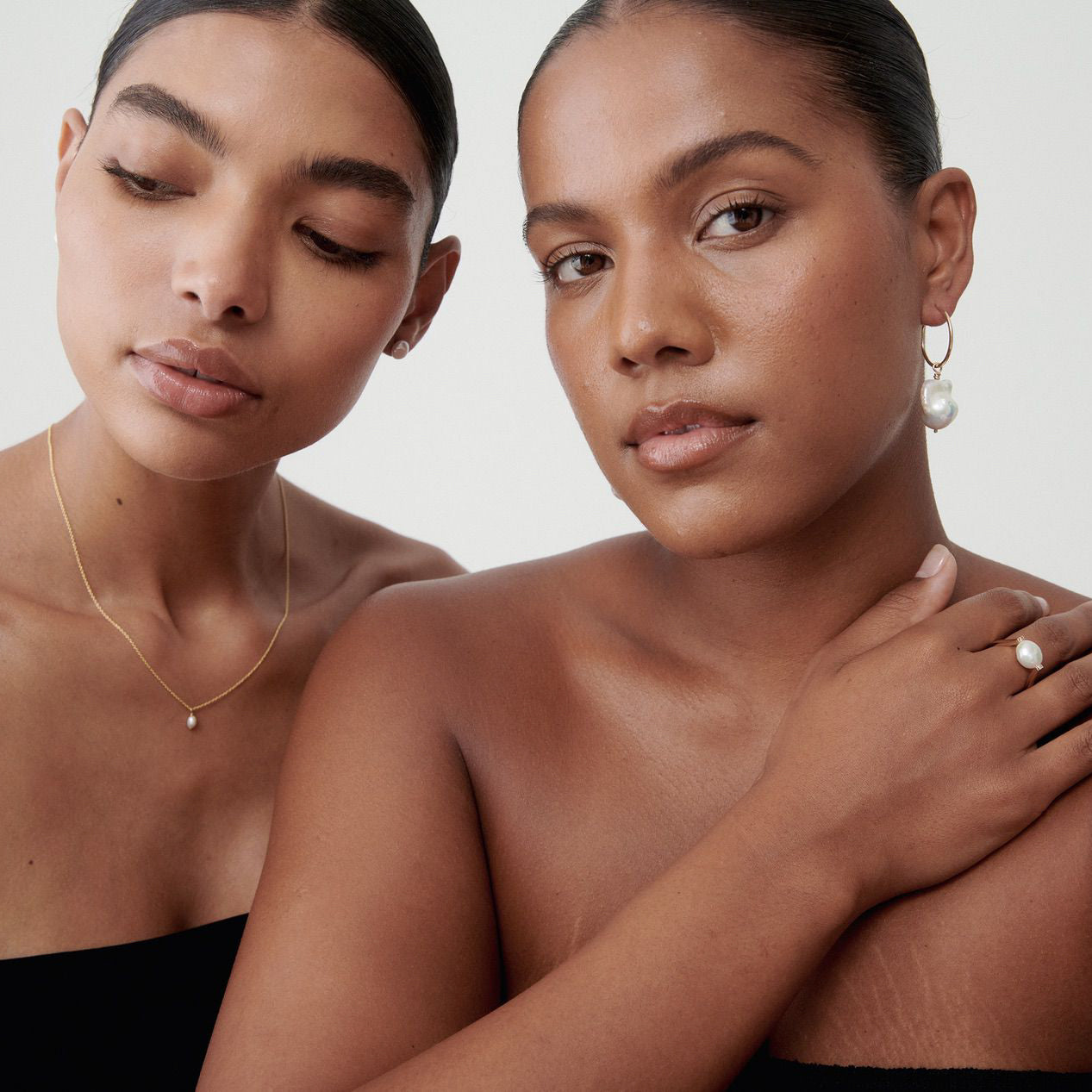 Two women showcase luxury jewellery. One wears a gold necklace with a pearl, and the other dons pearl earrings and a matching ring.