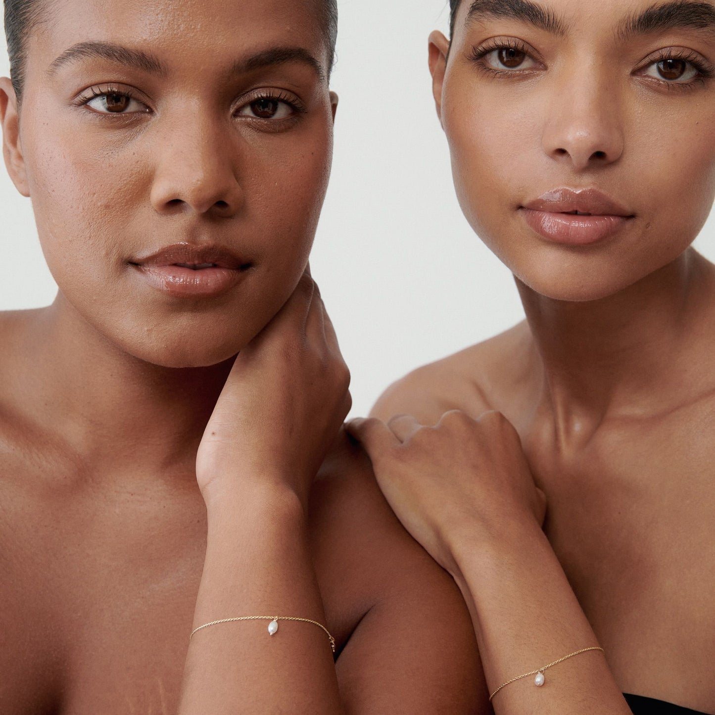 Two women pose gracefully, showcasing delicate gold bracelets with small pendants. Minimalist luxury jewellery style.
