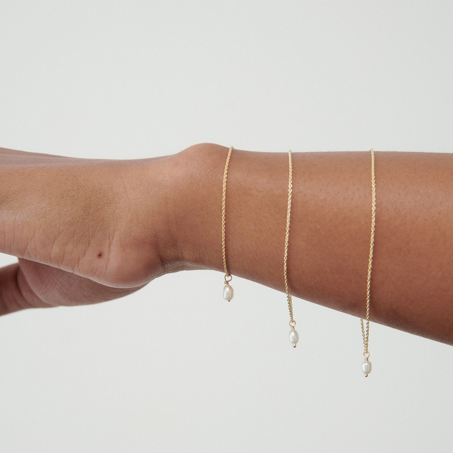 Three gold chain bracelets with small pearls elegantly draped on an outstretched arm against a pale background.